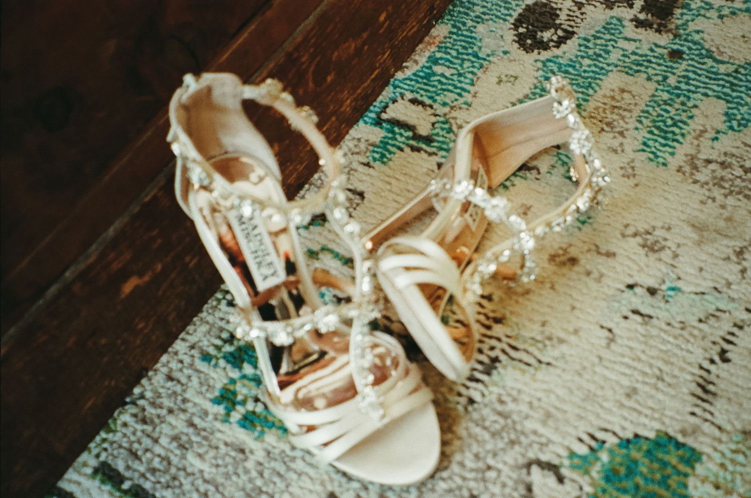 Elegant white bridal heeled sandals adorned with pearl embellishments displayed on a wooden floor with a teal rug.