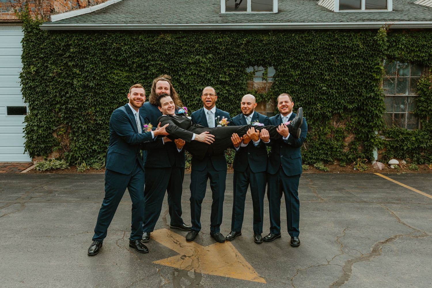 Groom lifted horizontally by six laughing groomsmen in navy suits in outdoor parking area. Chicagoland wedding venues.