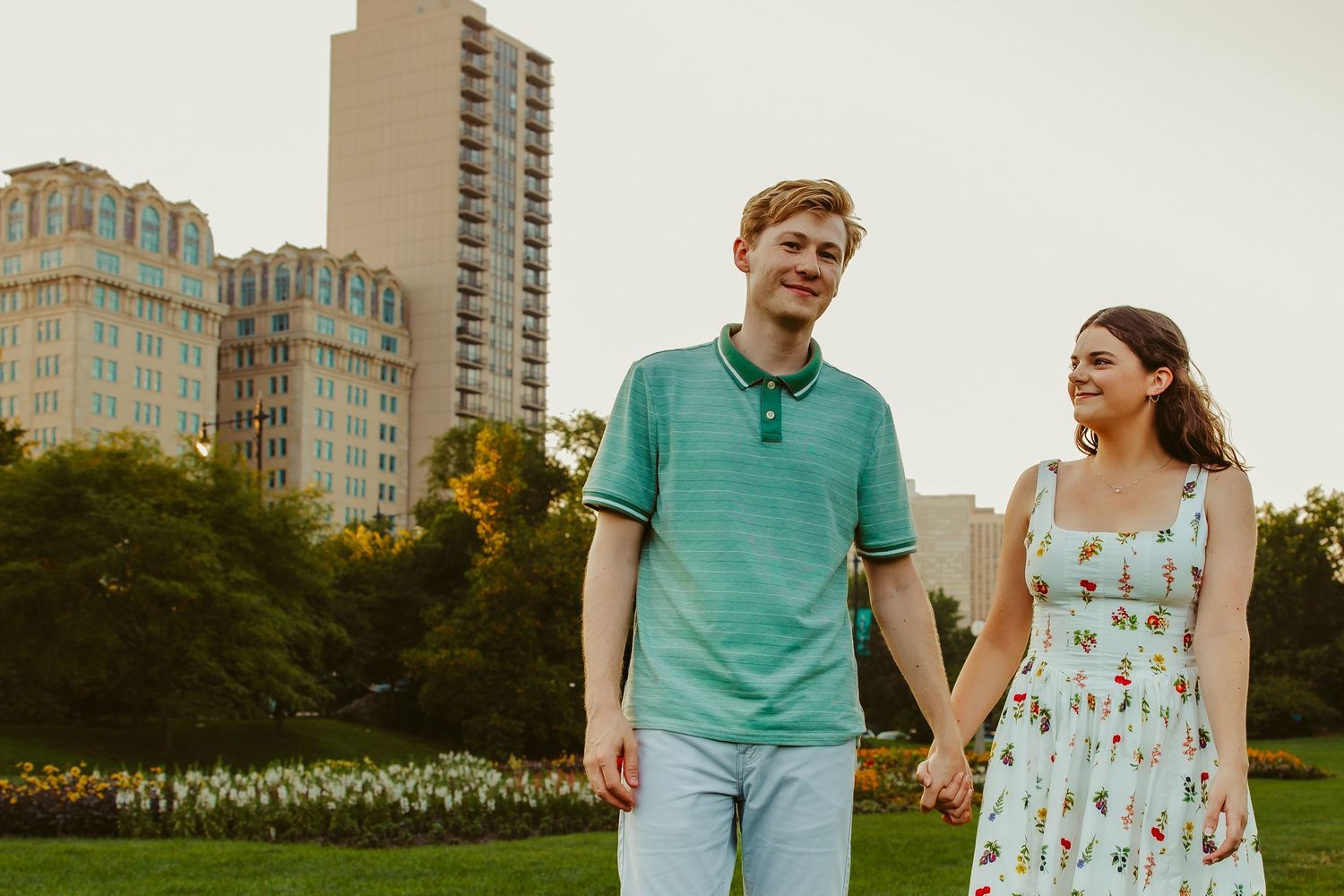Smiling couple holding hands in a park with colorful flower beds and urban buildings in the background. Chicago engagement photos.