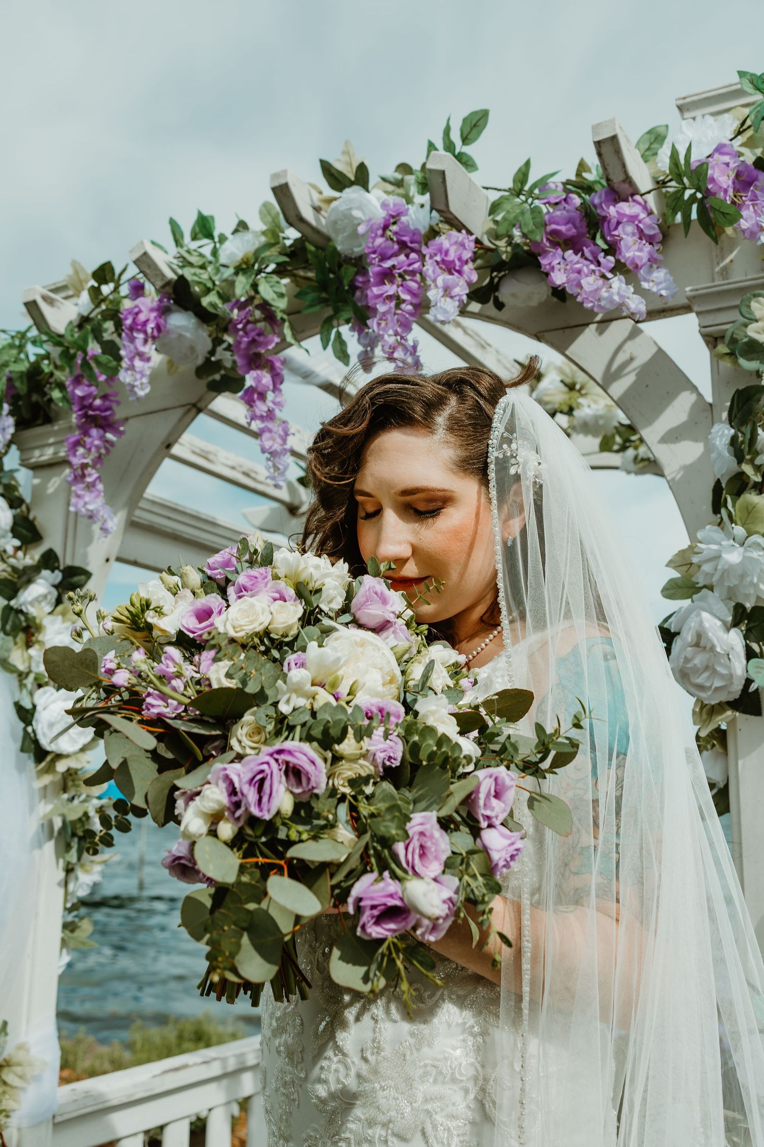 Bride in lace wedding gown holds lush purple and white bouquet under floral arch at lakeside ceremony.