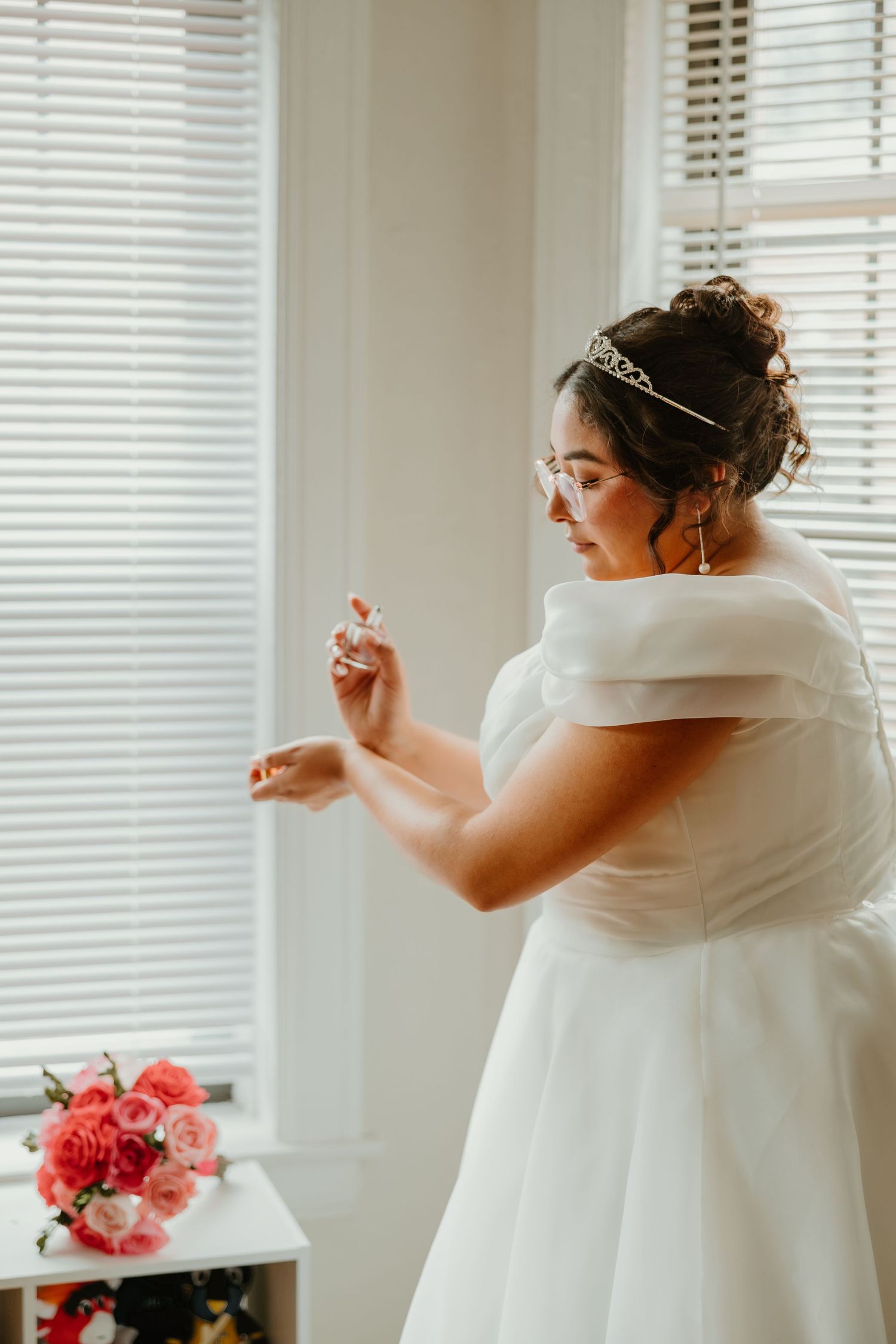 Bride in off-shoulder white gown applies perfume by window, wearing tiara and glasses with pink bouquet nearby. Chicagoland weddings.