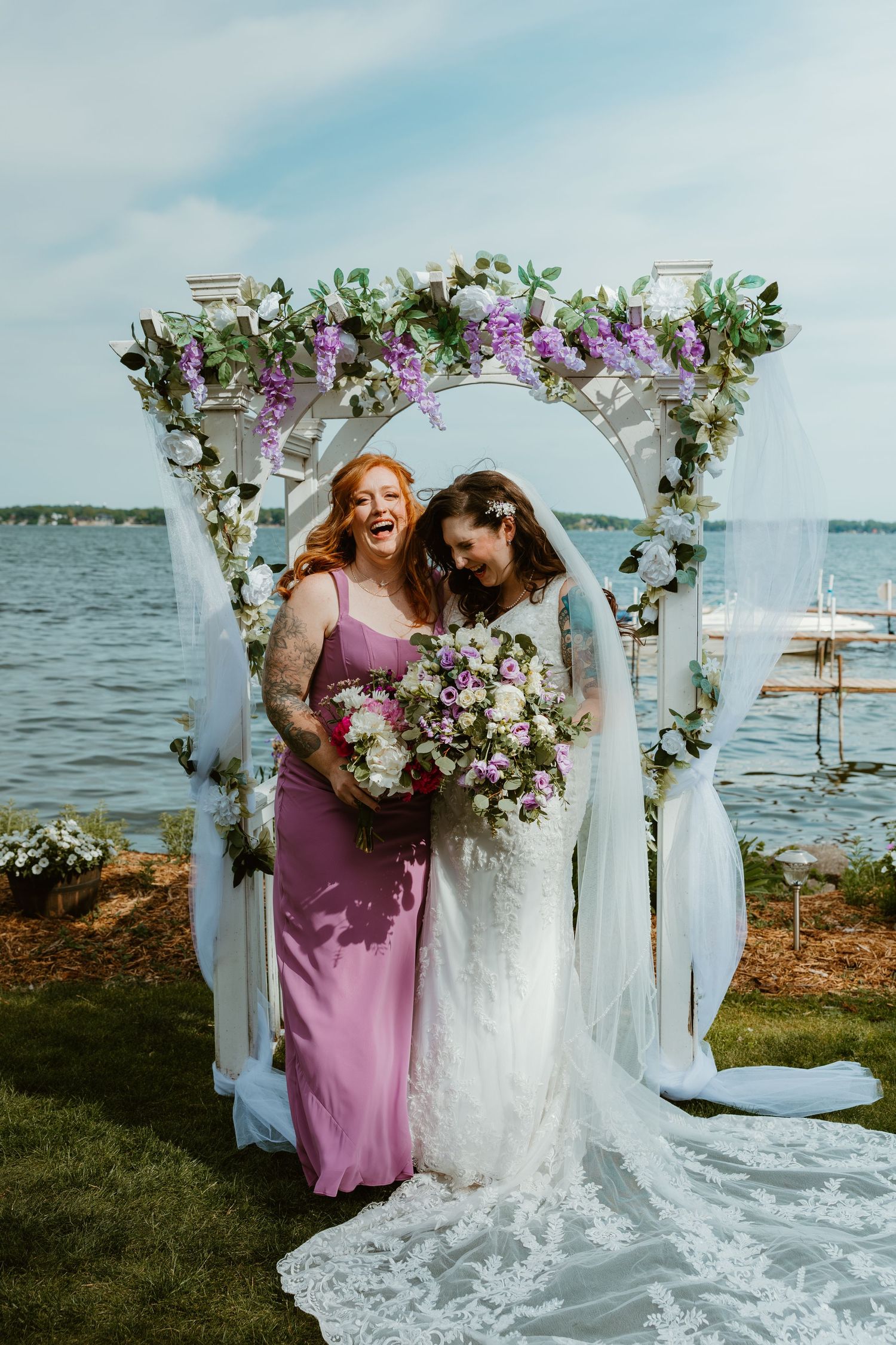 Bride and bridesmaid in purple dress laugh joyfully together under a floral arch by the lake.