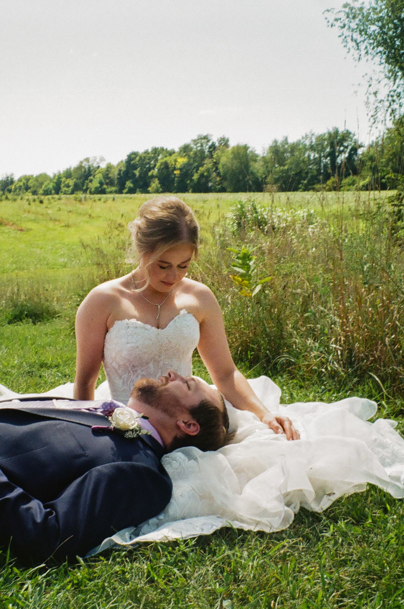 Bride in white lace gown sits on grass as groom in navy suit lies on her dress in a lush green field.