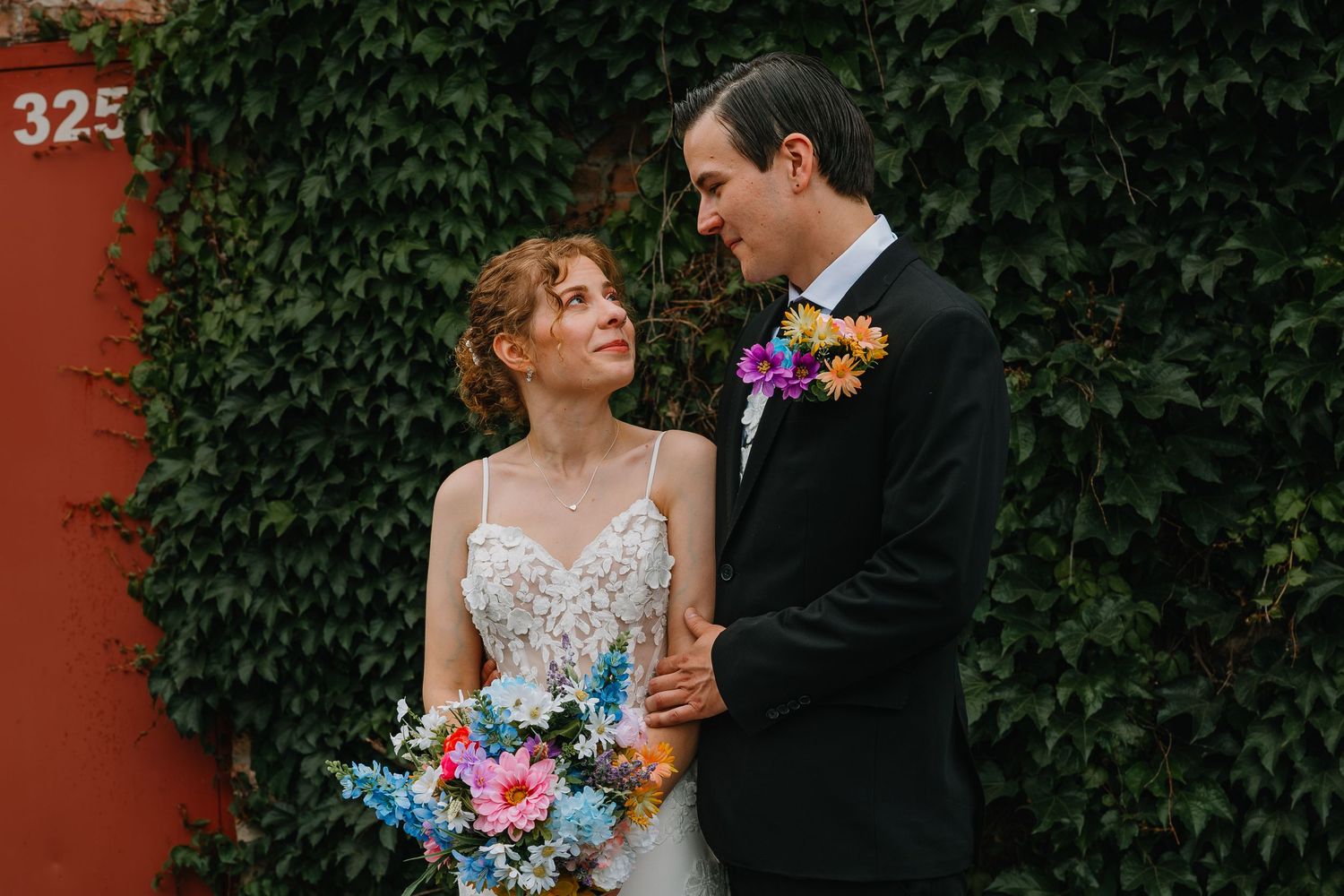 Bride in lace gown holding colorful bouquet gazes at groom in black tux against ivy-covered red wall. Chicagoland wedding venues.