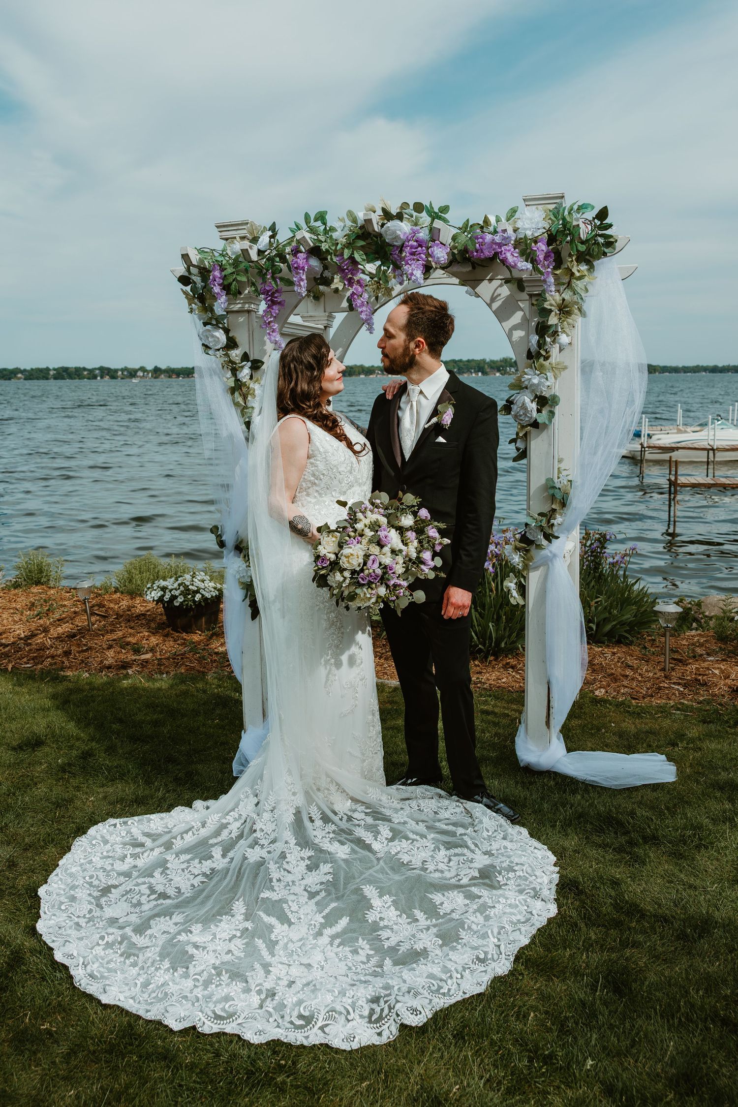 Bride in lace gown with long train and groom pose under white floral arch beside serene lake.