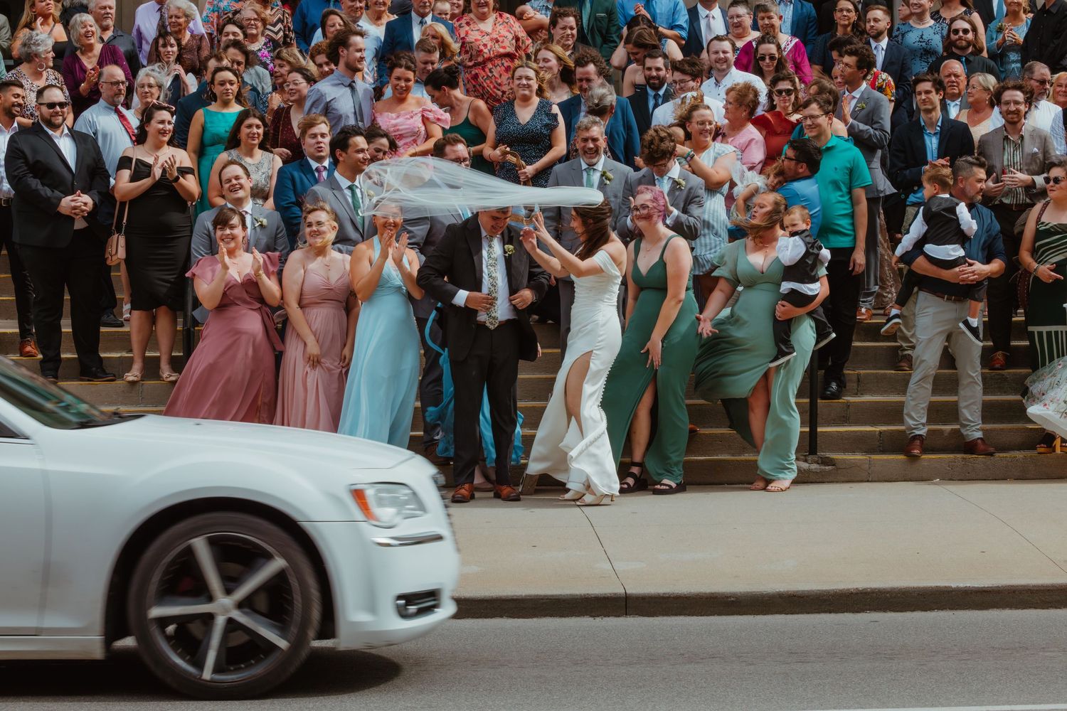 Wedding party and guests celebrating outside a church as a white car passes on the street.