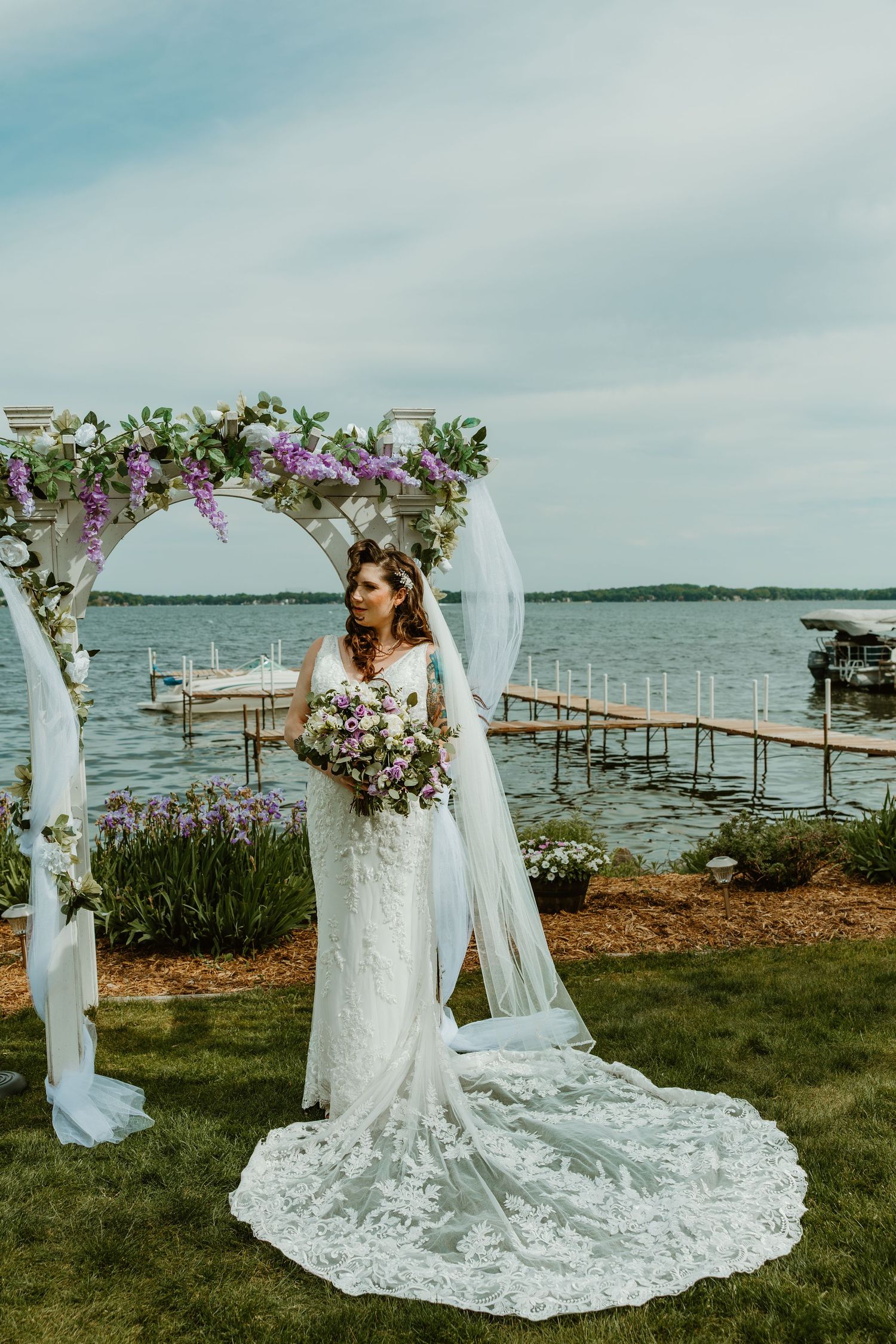 Bride in lace gown with long train poses under a floral arch with purple flowers by a lake.