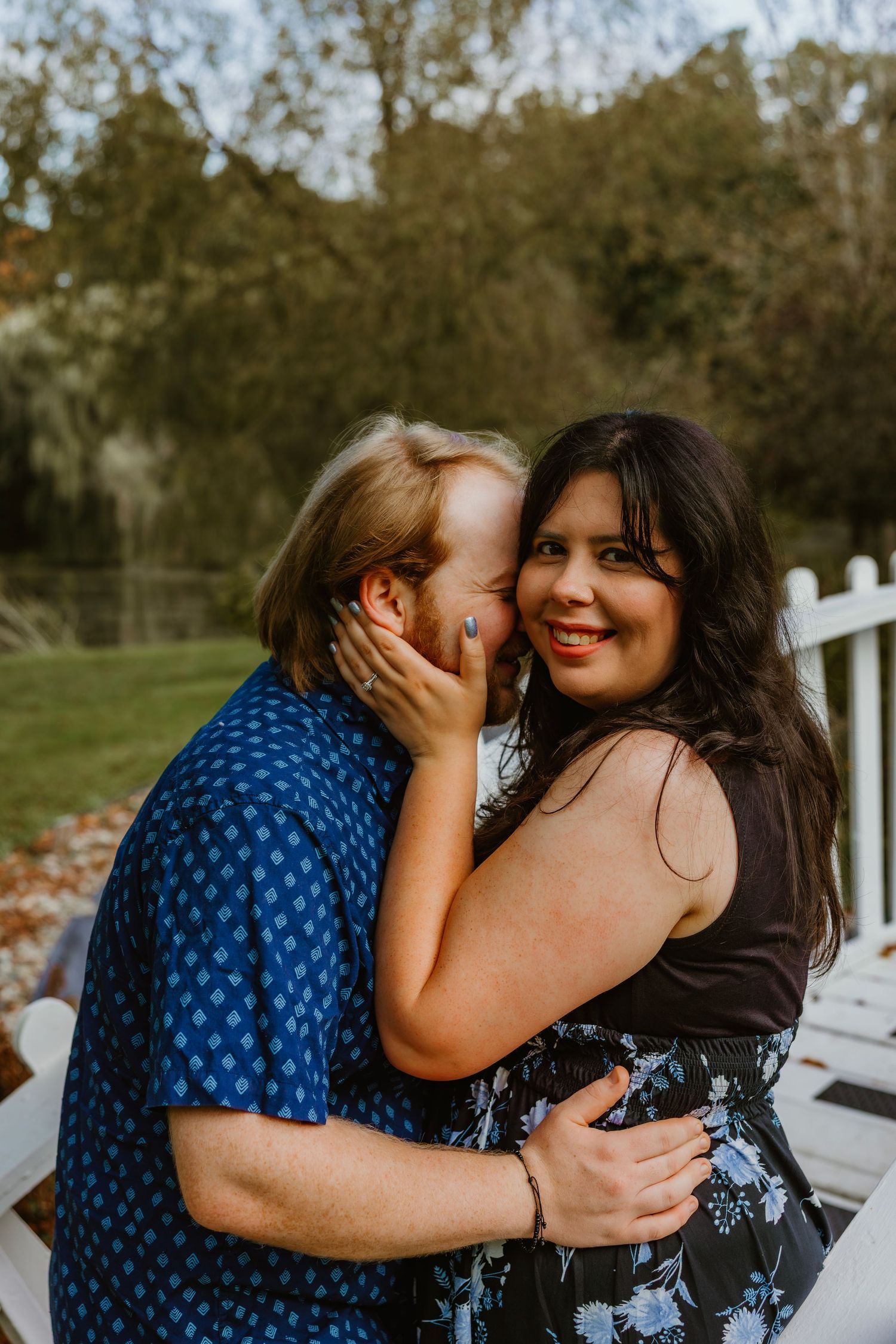 Affectionate couple embracing and smiling on a porch with autumn trees in the background. Northwest Indiana wedding engagement.