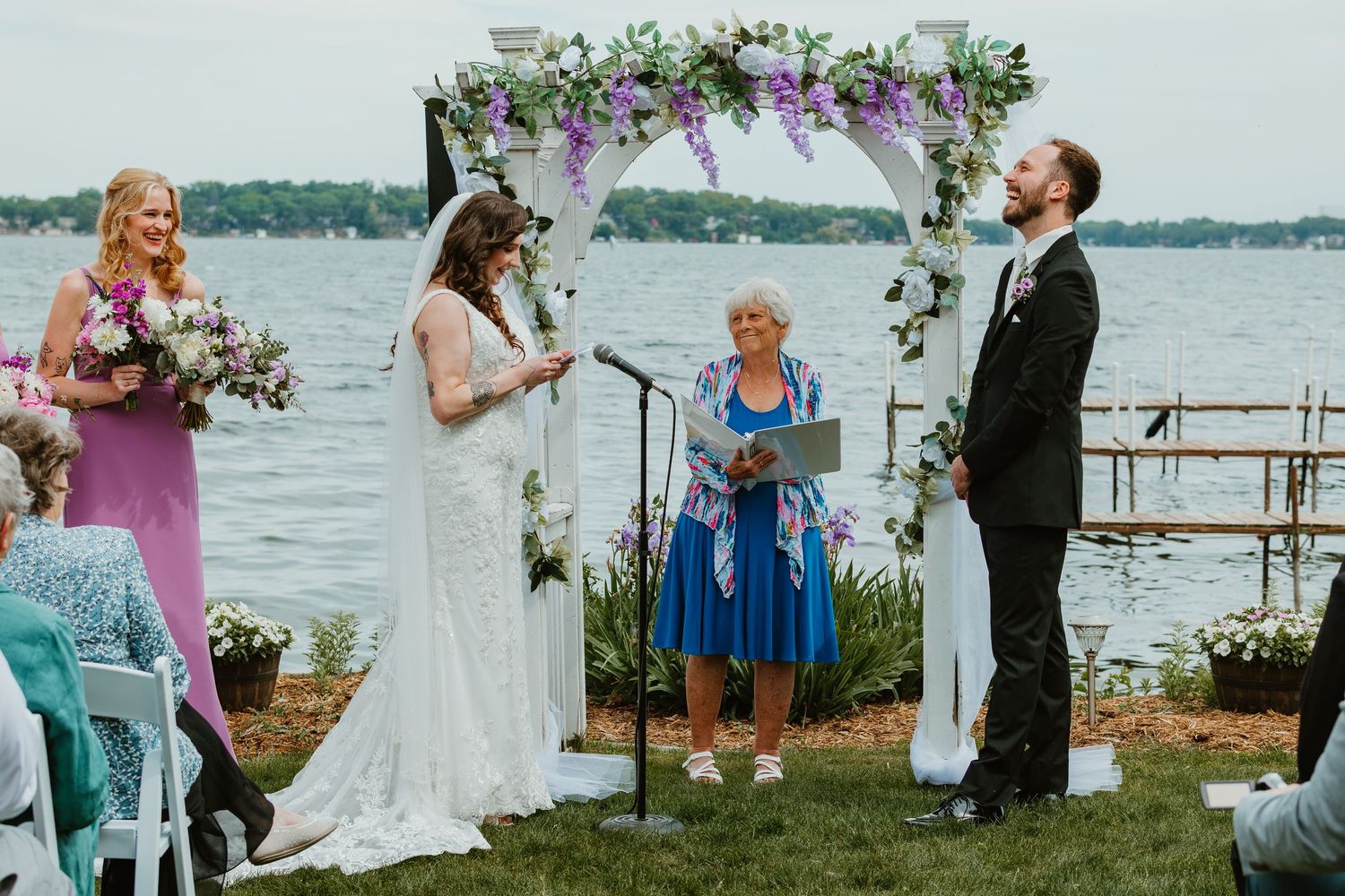 Bride and groom exchange vows under floral arch by lakeside with officiant and bridesmaid during outdoor wedding ceremony.
