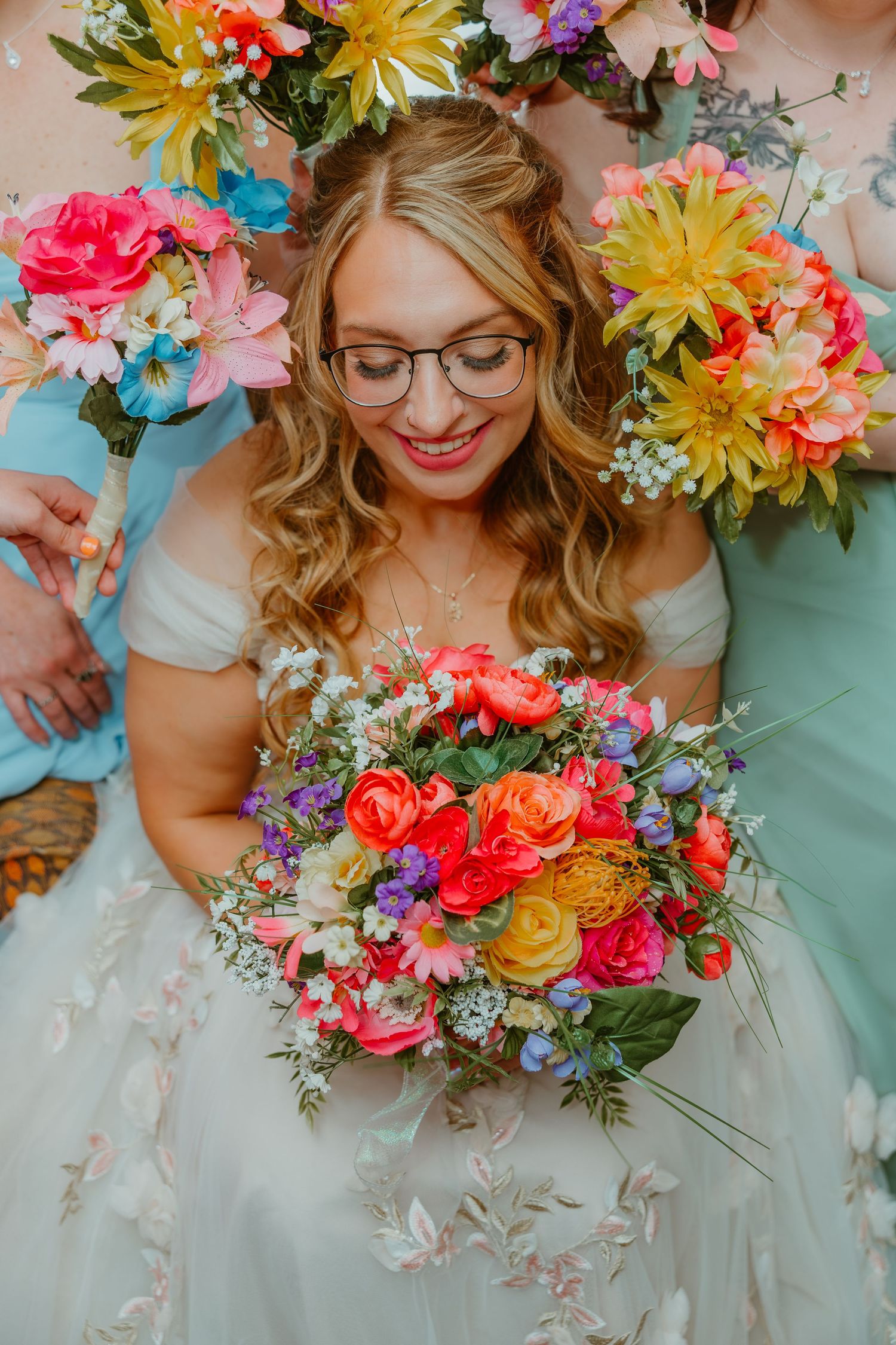 Smiling bride with glasses holds vibrant multicolored bouquet surrounded by colorful floral arrangements.