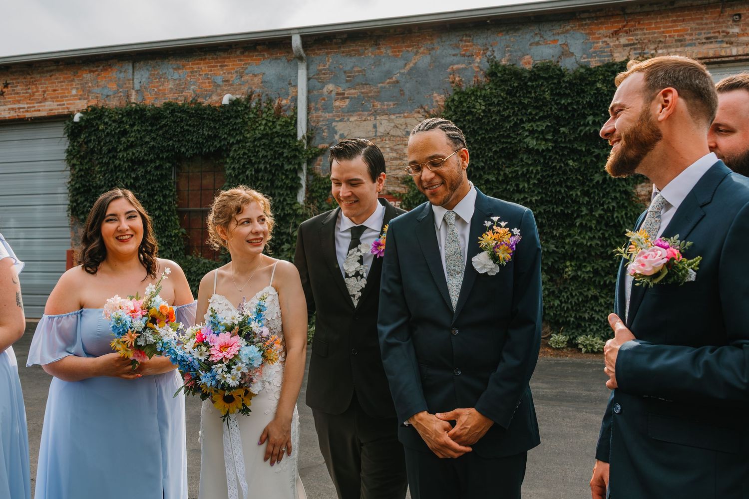Bride with colorful bouquet stands with groom and wedding party smiling outside rustic brick building. Chicagoland wedding venues.