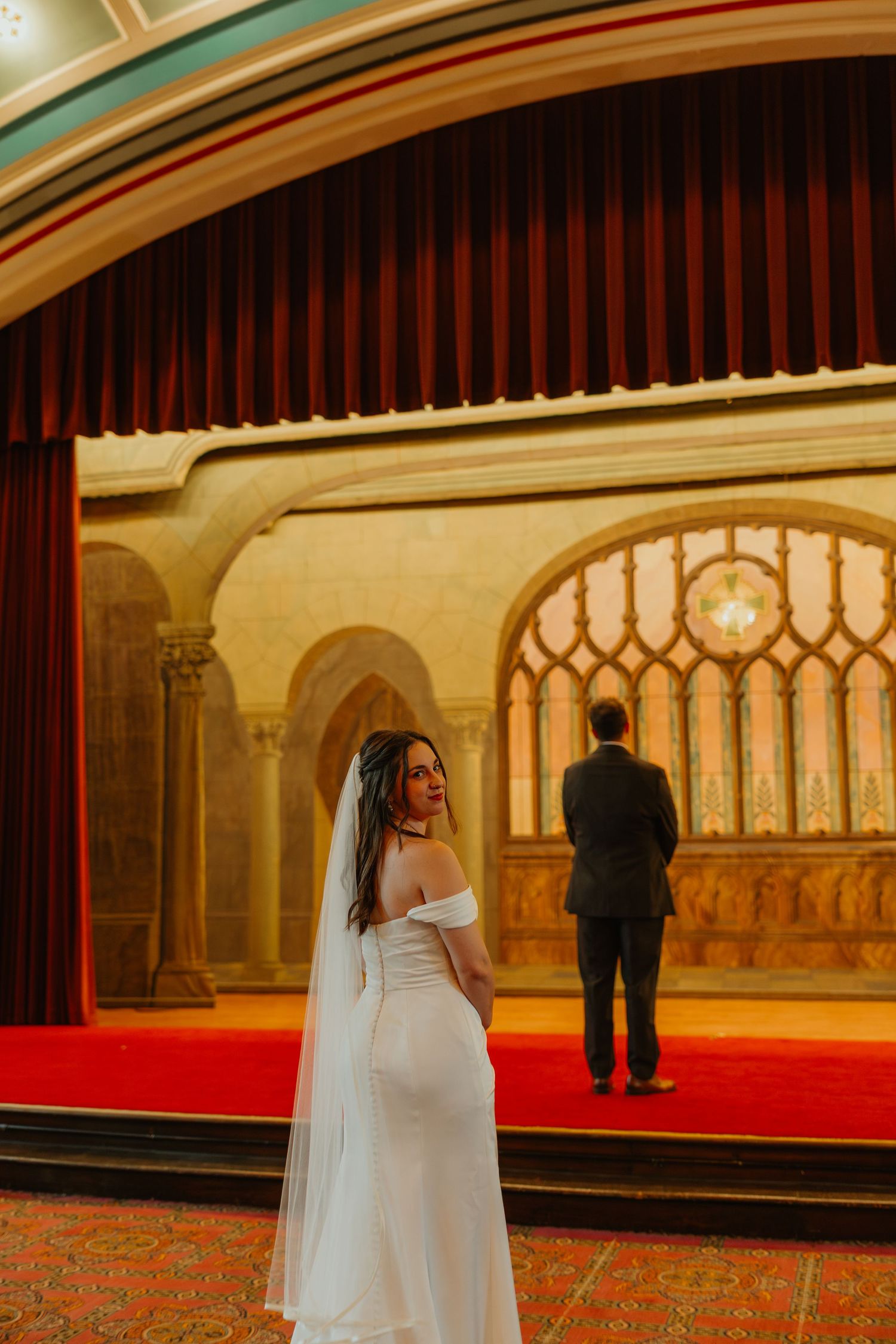 Bride in white off-shoulder gown with veil posing in front of an ornate theater stage backdrop.