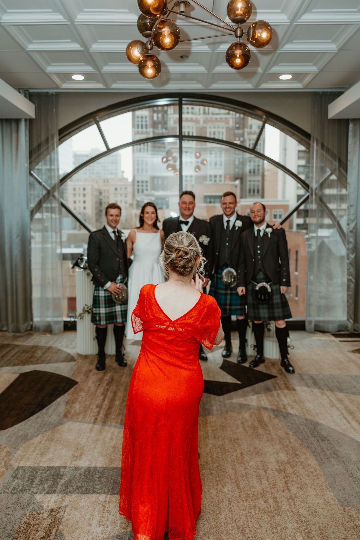 Wedding party in kilts posing under an arched window as a woman in a red gown faces them. Chicago Thompson Hotel wedding.