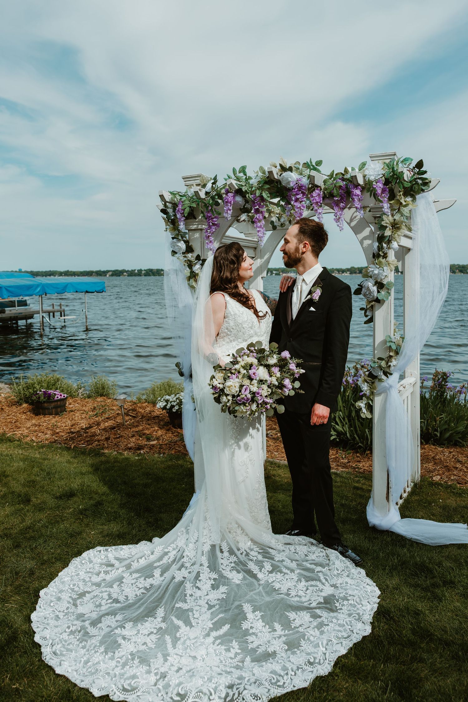 Bride and groom share a romantic moment under a floral arch adorned with purple wisteria by a lakeside.
