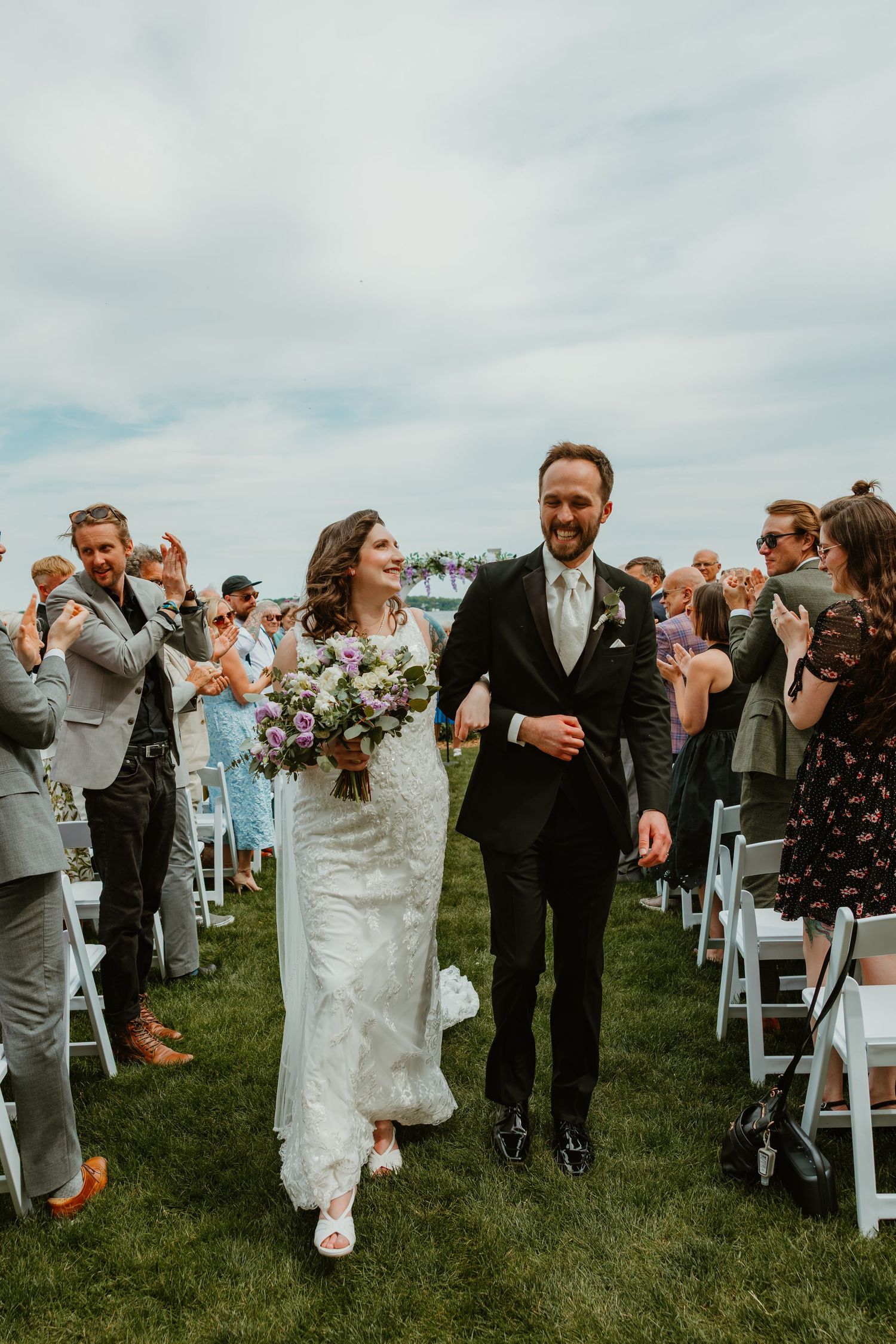 Newlywed couple walks down outdoor wedding aisle as guests applaud, bride holding purple bouquet in lace gown.