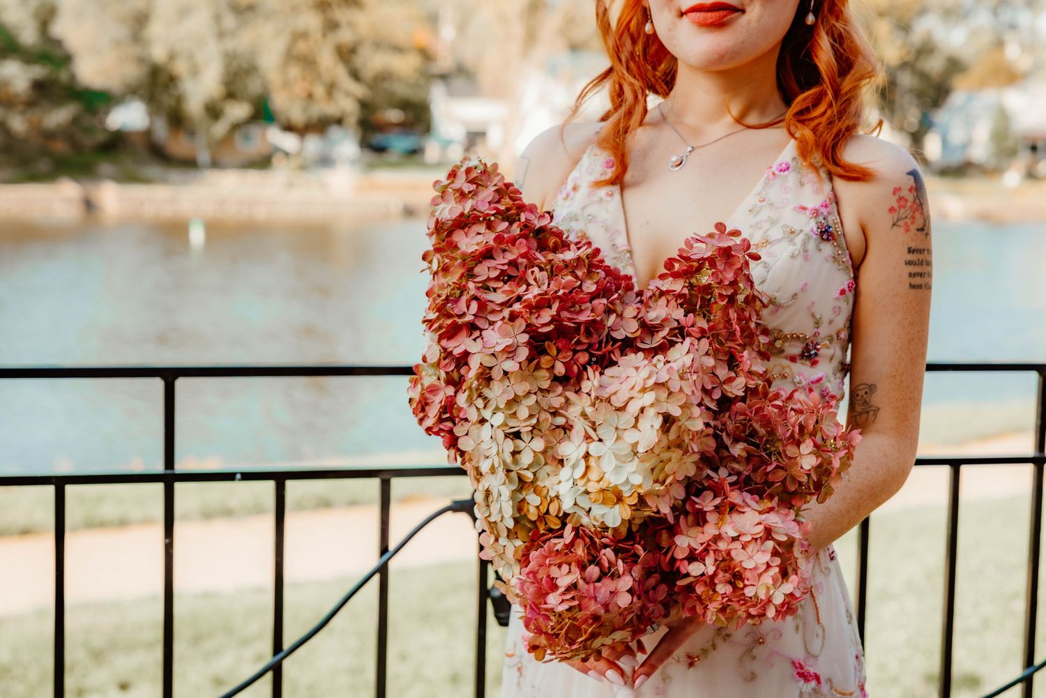 Red-haired bride holding a large dried hydrangea bouquet by a waterfront railing.
