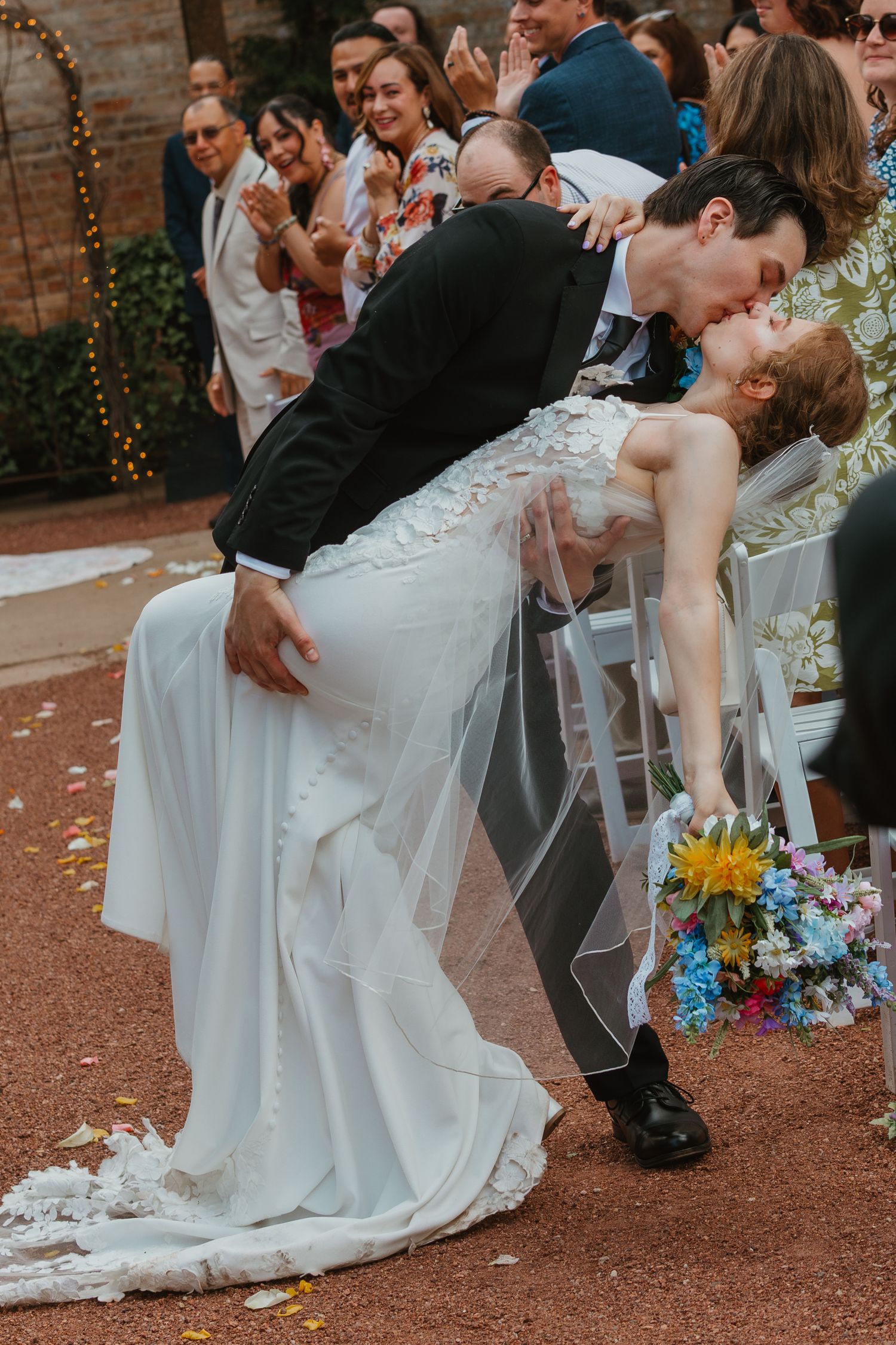 Groom dips bride for romantic kiss after outdoor wedding ceremony while guests celebrate with colorful confetti. Chicagoland wedding venues.
