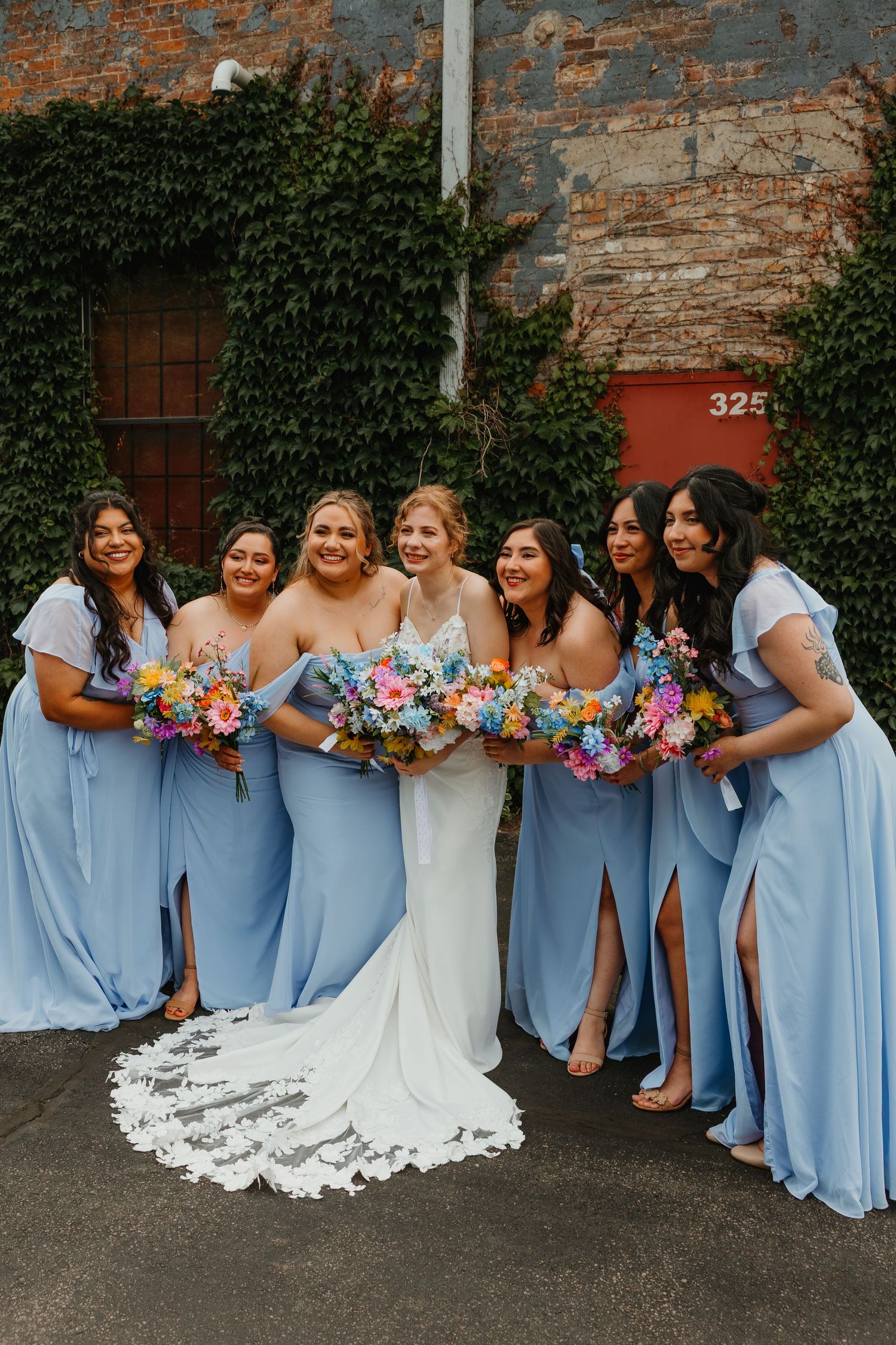 Bride in lace gown poses with six bridesmaids in light blue dresses holding colorful bouquets. Chicagoland wedding venues.