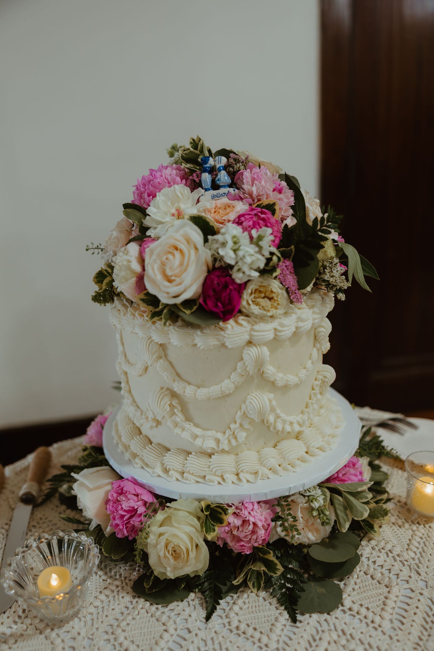 Two-tier white wedding cake adorned with pink peonies, cream roses, and lush greenery on lace tablecloth.