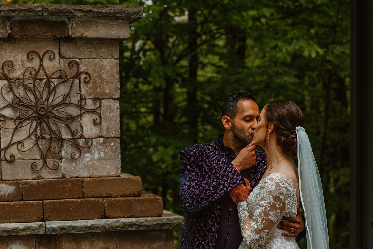 Groom tenderly kissing bride near a decorative stone fireplace in a woodland wedding setting. Documentary style wedding photos.