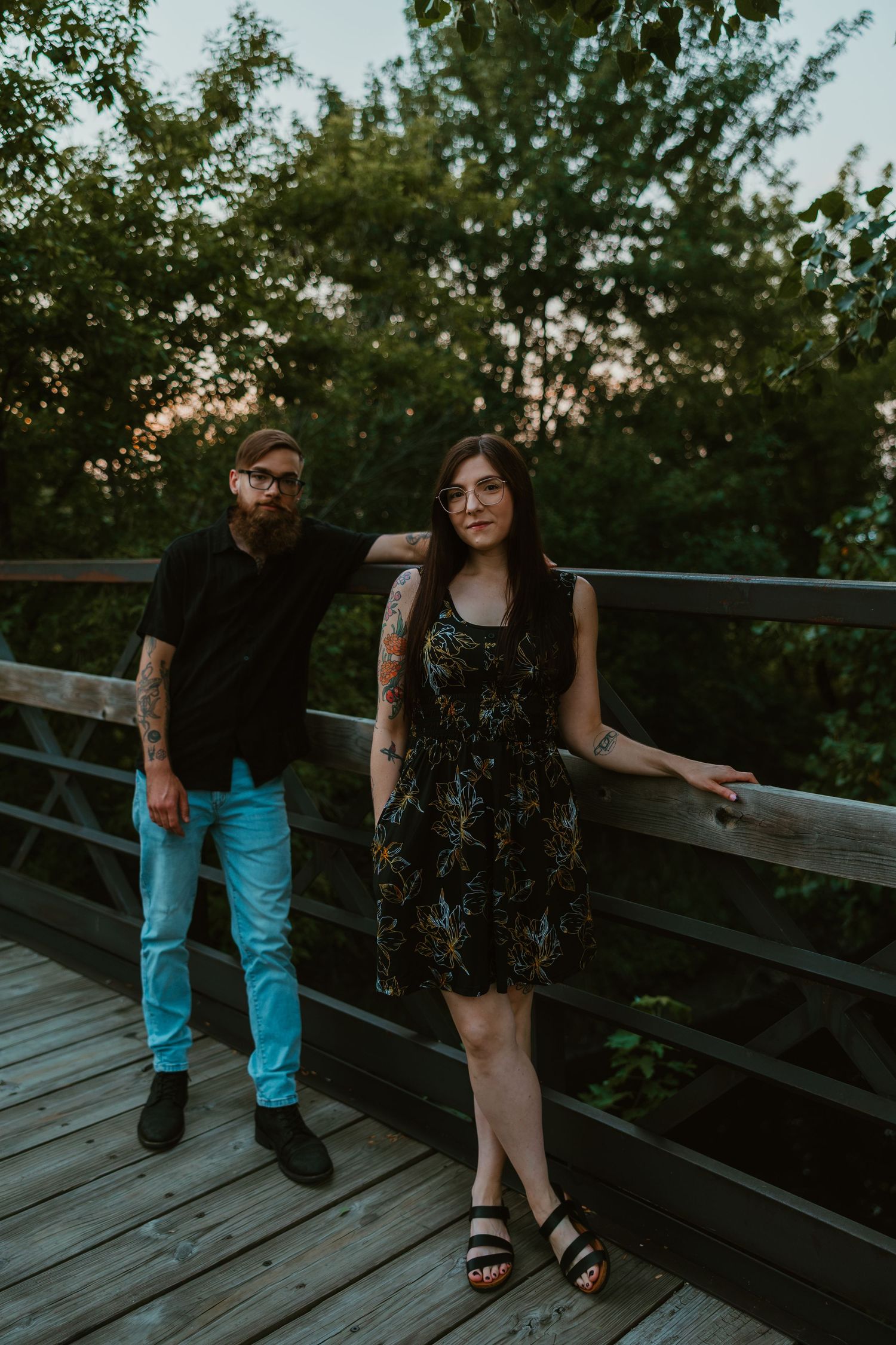 Tattooed couple posing on a wooden bridge at golden hour, surrounded by green trees in summer. Chicagoland engagement photos.