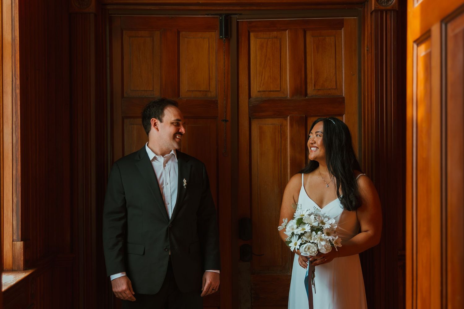 Smiling bride with white bouquet and groom share a loving glance by elegant wooden church doors. Michigan elopement.
