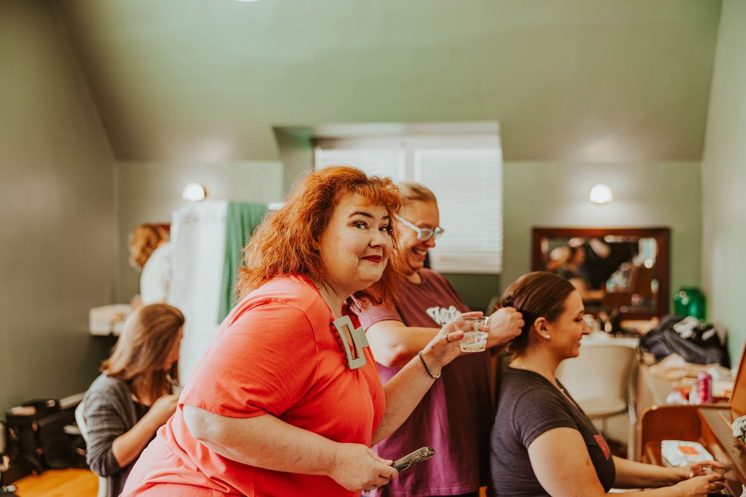 Hair stylist with red curly hair works on bridesmaid's hair in busy wedding preparation room.