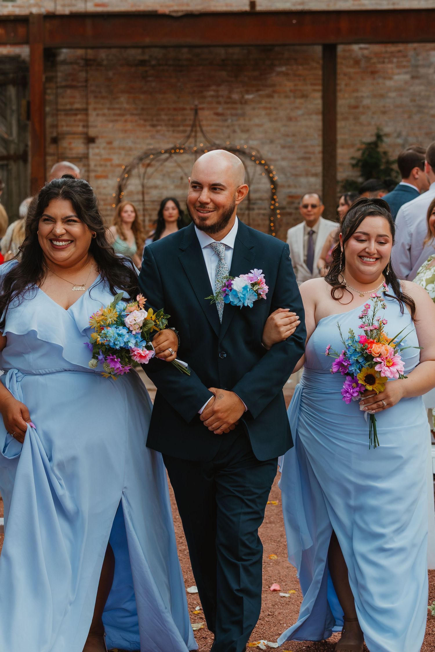 A groom in a navy suit walks down the aisle with two bridesmaids in light blue gowns holding bouquets. Chicagoland wedding venues.