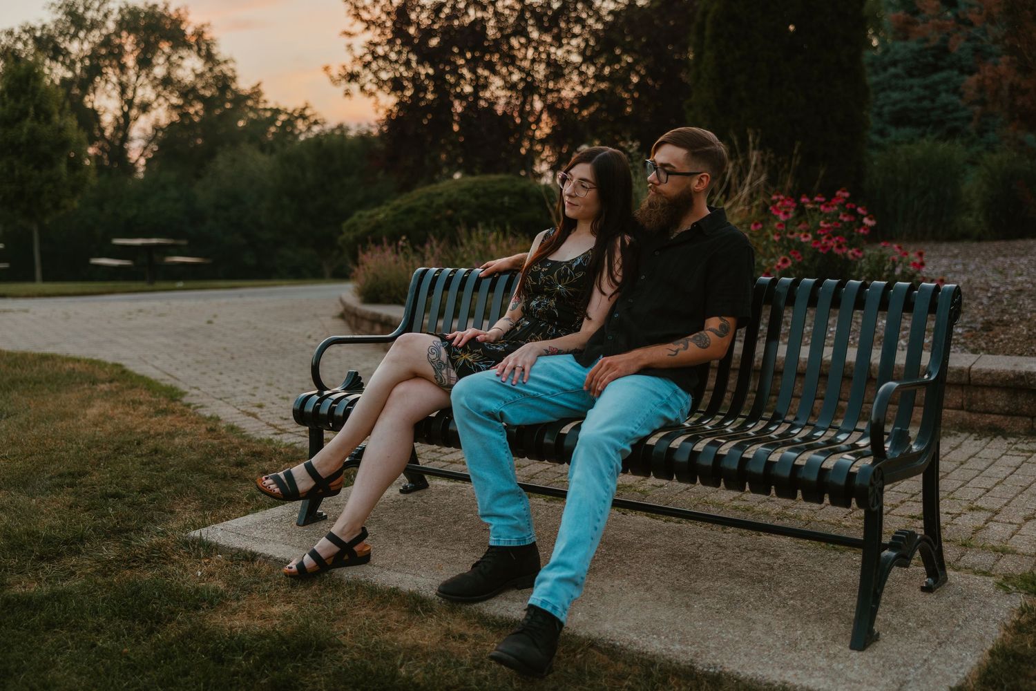 Tattooed couple sitting on a green park bench at sunset, surrounded by lush garden greenery. Chicagoland engagement photos.