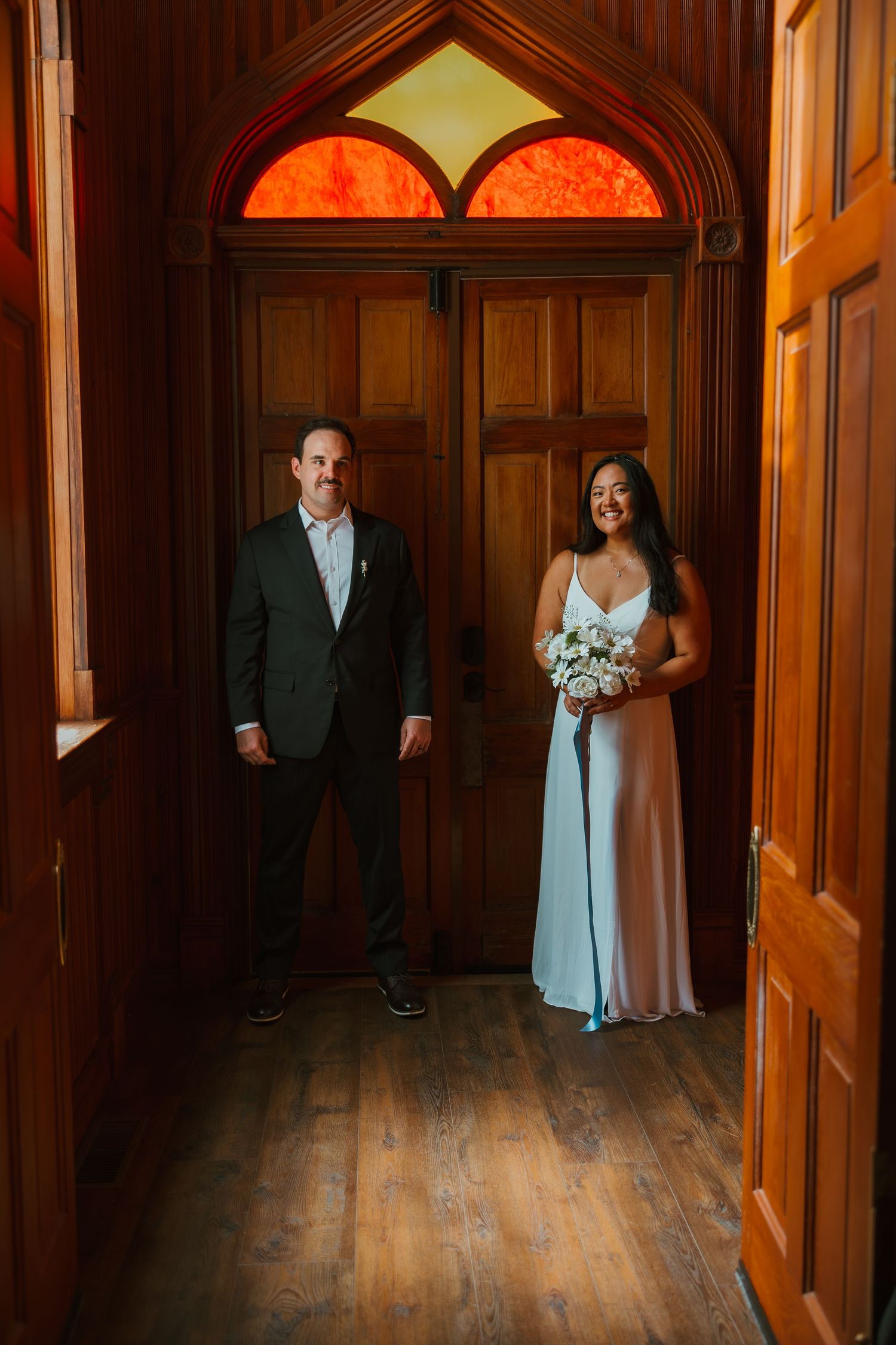 Bride holding white bouquet and groom standing apart in wooden church doorway with stained glass. Michigan elopement.