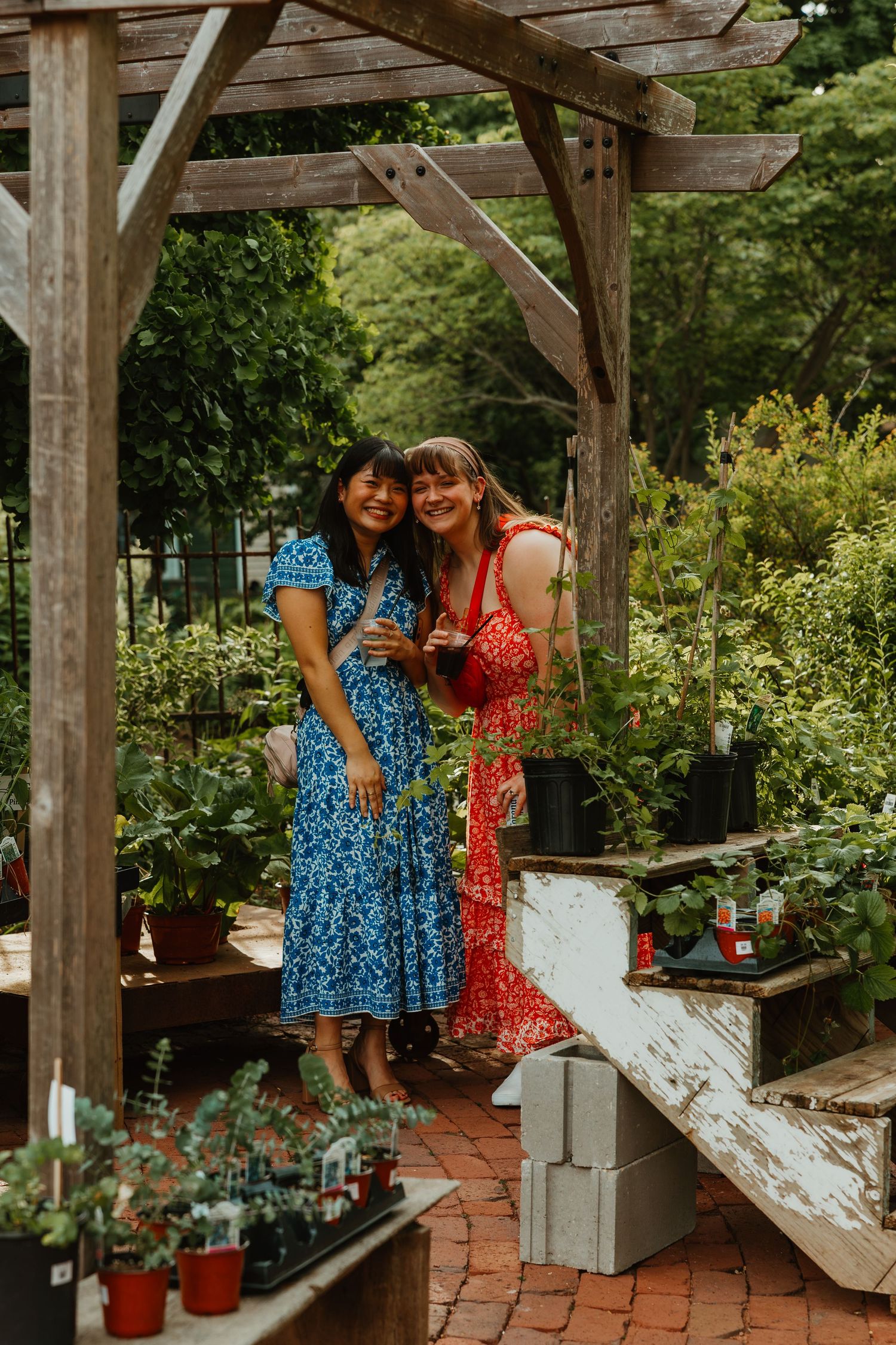 Two women in floral dresses smile together under a wooden pergola surrounded by lush greenery and potted plants. Chicagoland wedding venues.