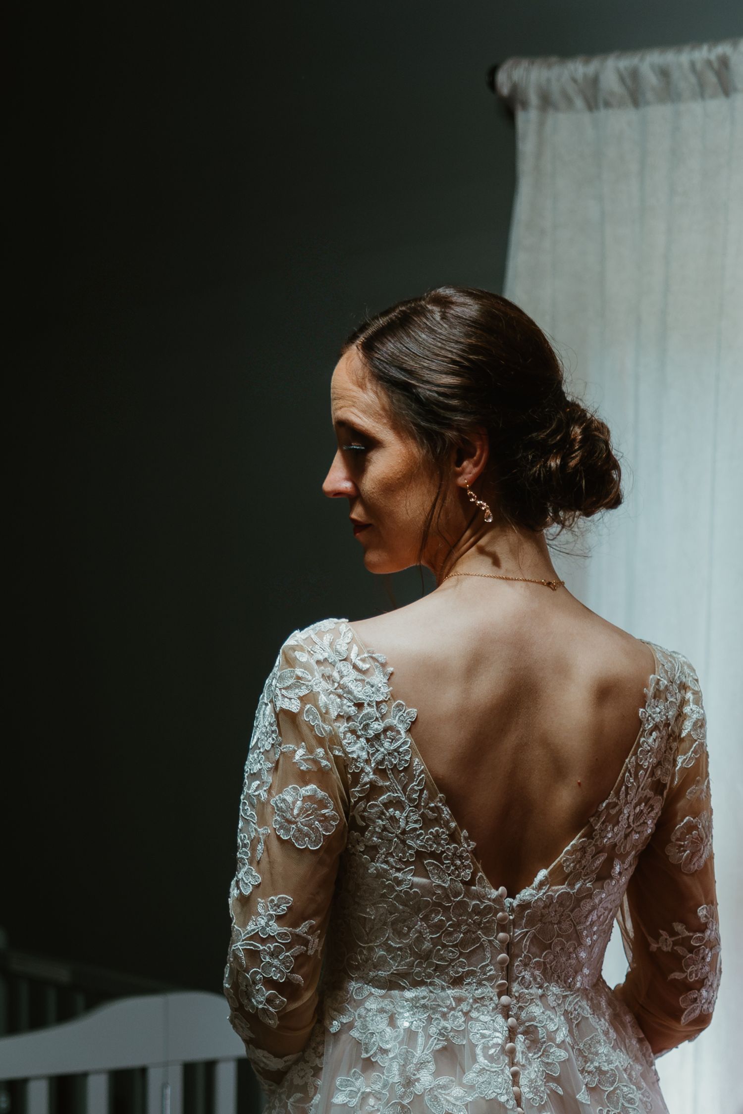 Bride in backless lace gown with updo stands by window in soft light. Documentary style wedding photos.