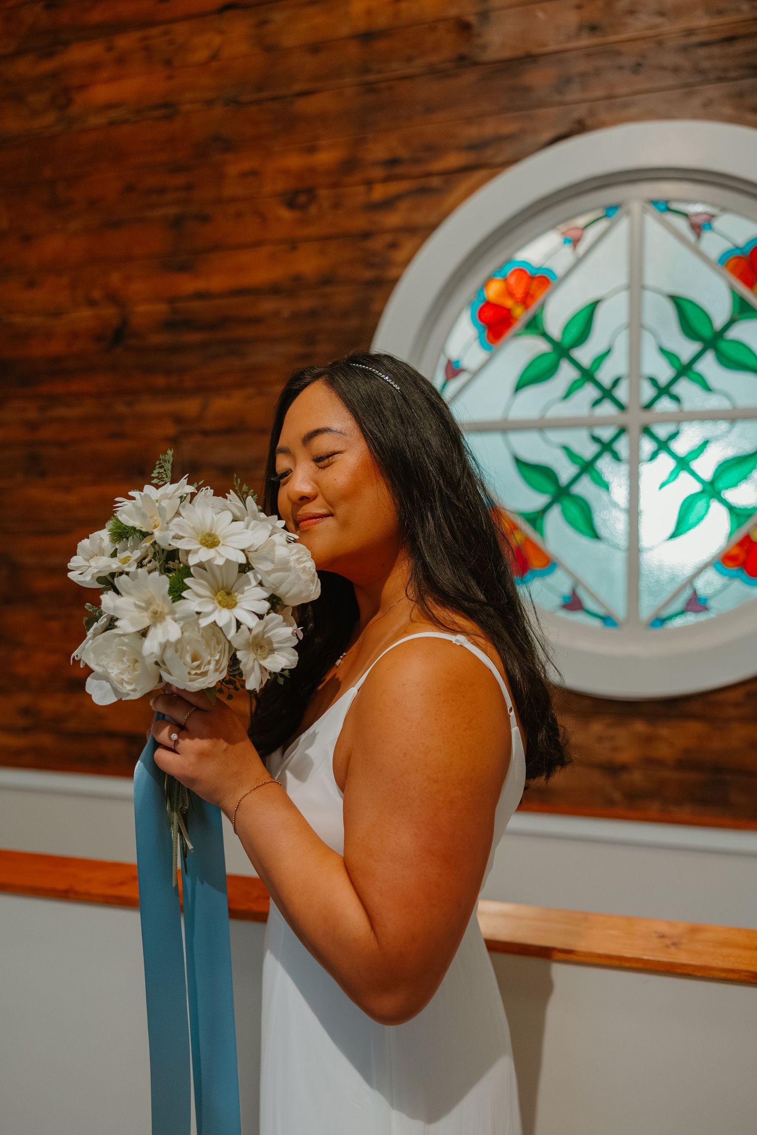 Smiling bride in white dress holds white daisy bouquet with blue ribbon near stained glass window. Michigan elopement.