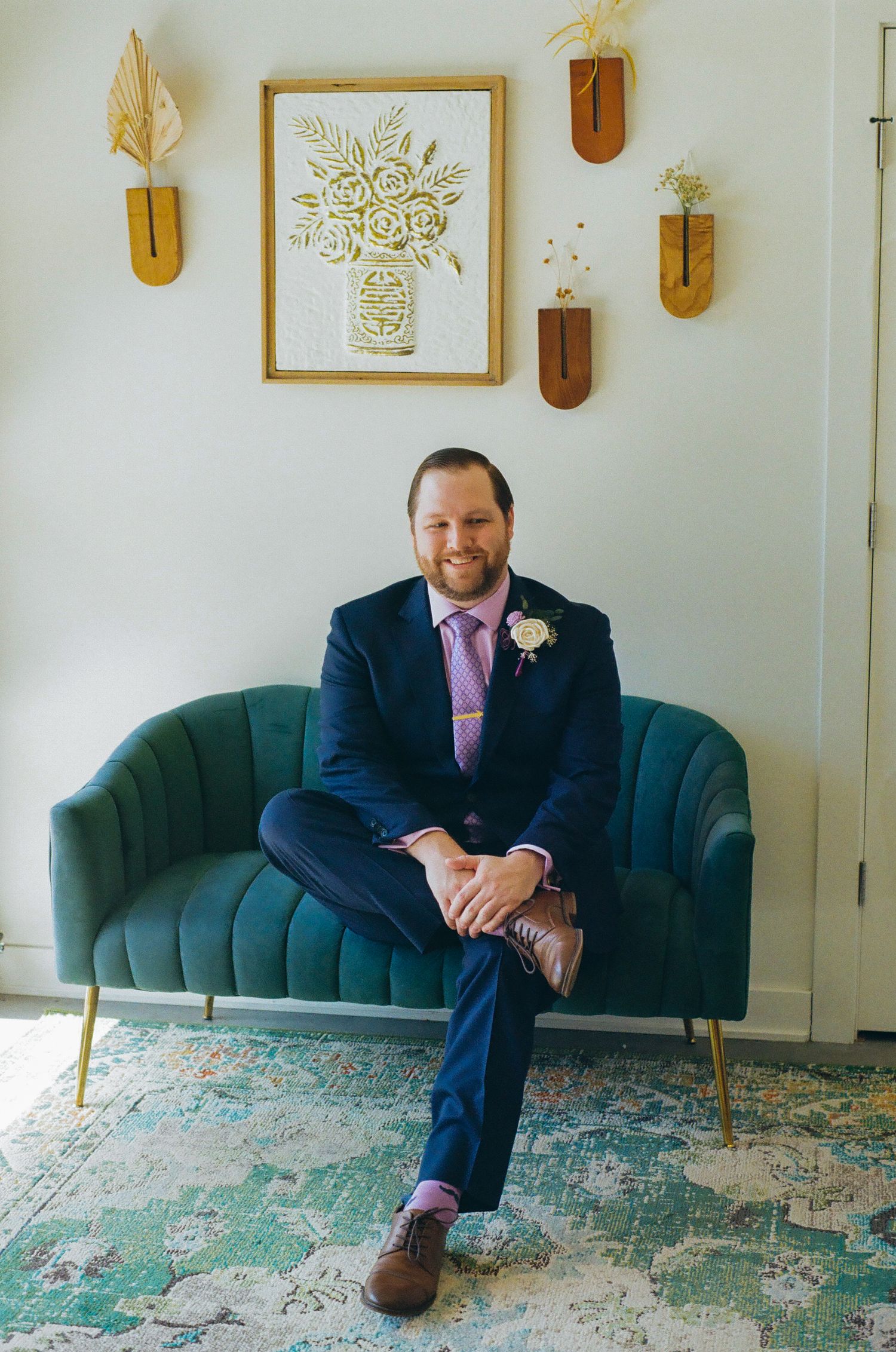 A smiling groom in a navy suit with lavender tie and boutonniere sits on a teal velvet loveseat in an elegant room.