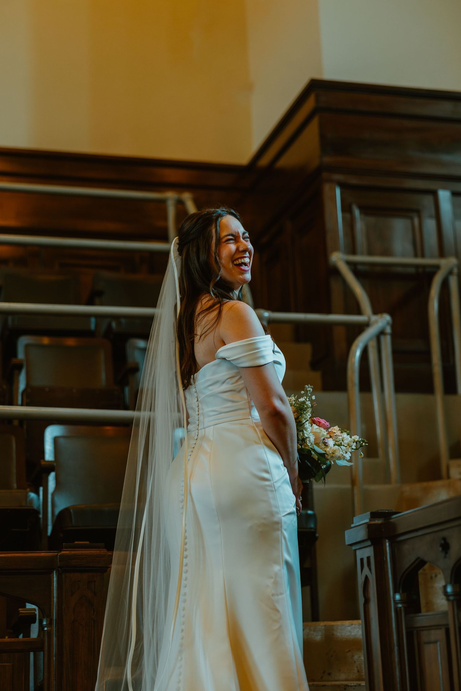 Radiant bride in off-shoulder satin gown with cathedral veil laughs joyfully on church staircase.