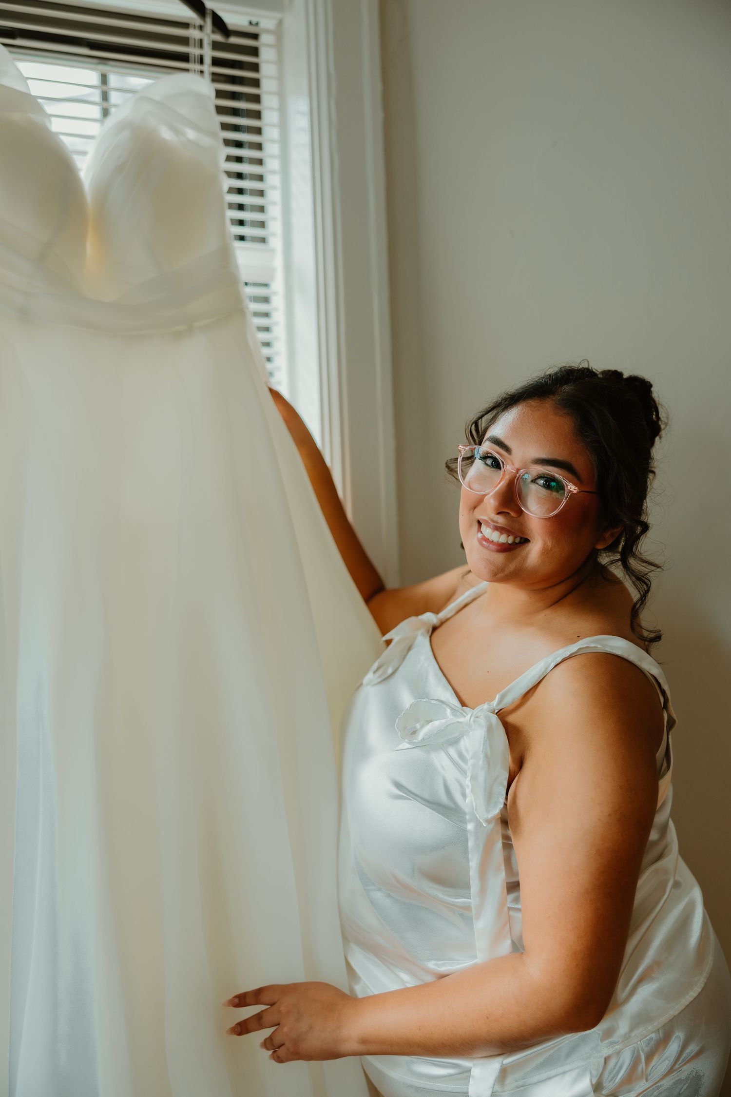 Excited bride in white satin romper and glasses smiles beside her strapless wedding gown hanging in window. Chicagoland weddings.