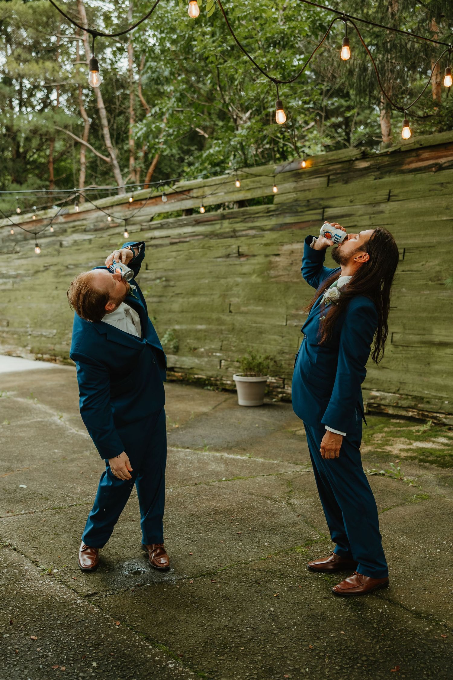 Two groomsmen in navy blue suits drinking cans outdoors at wedding reception near rustic wood wall. Northwest Indiana wedding venues.