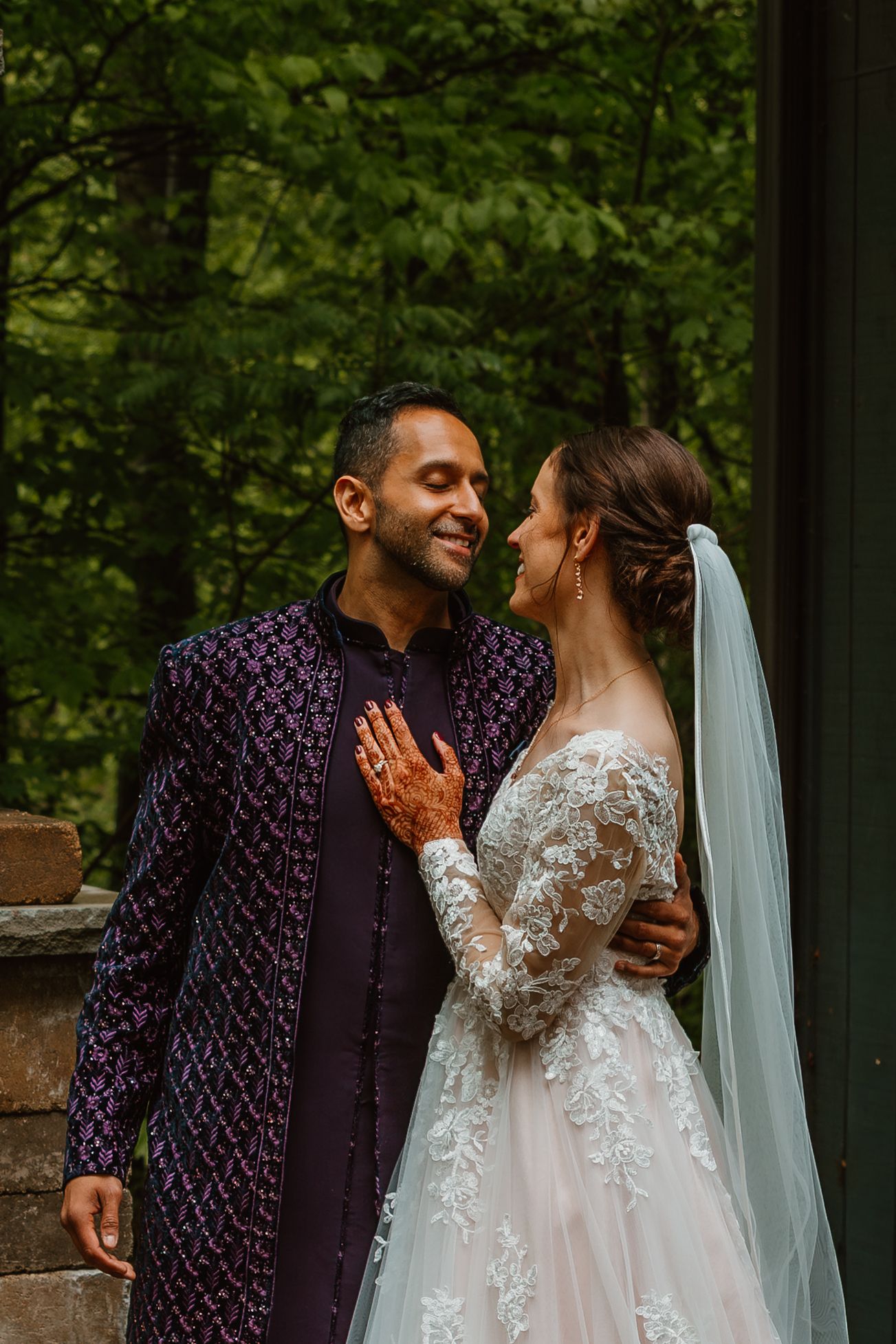 Groom in purple embroidered sherwani embraces bride in lace gown outdoors. Documentary style wedding photos.