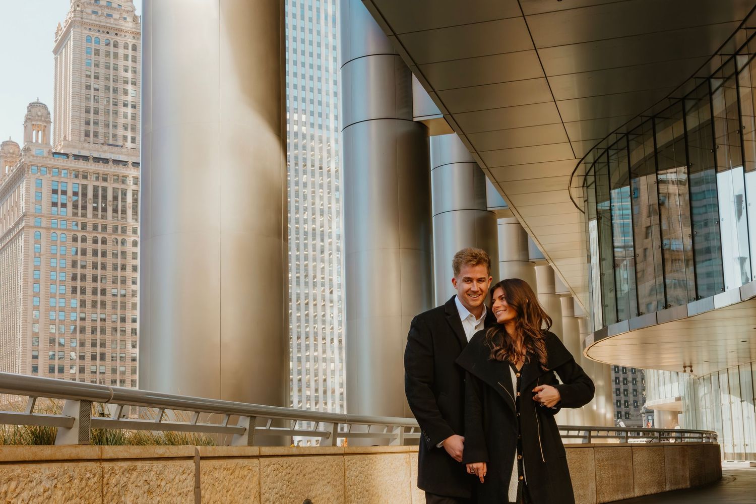 Engaged couple embracing near silver columns with Chicago skyline in background. Outdoor Chicago engagement photos.