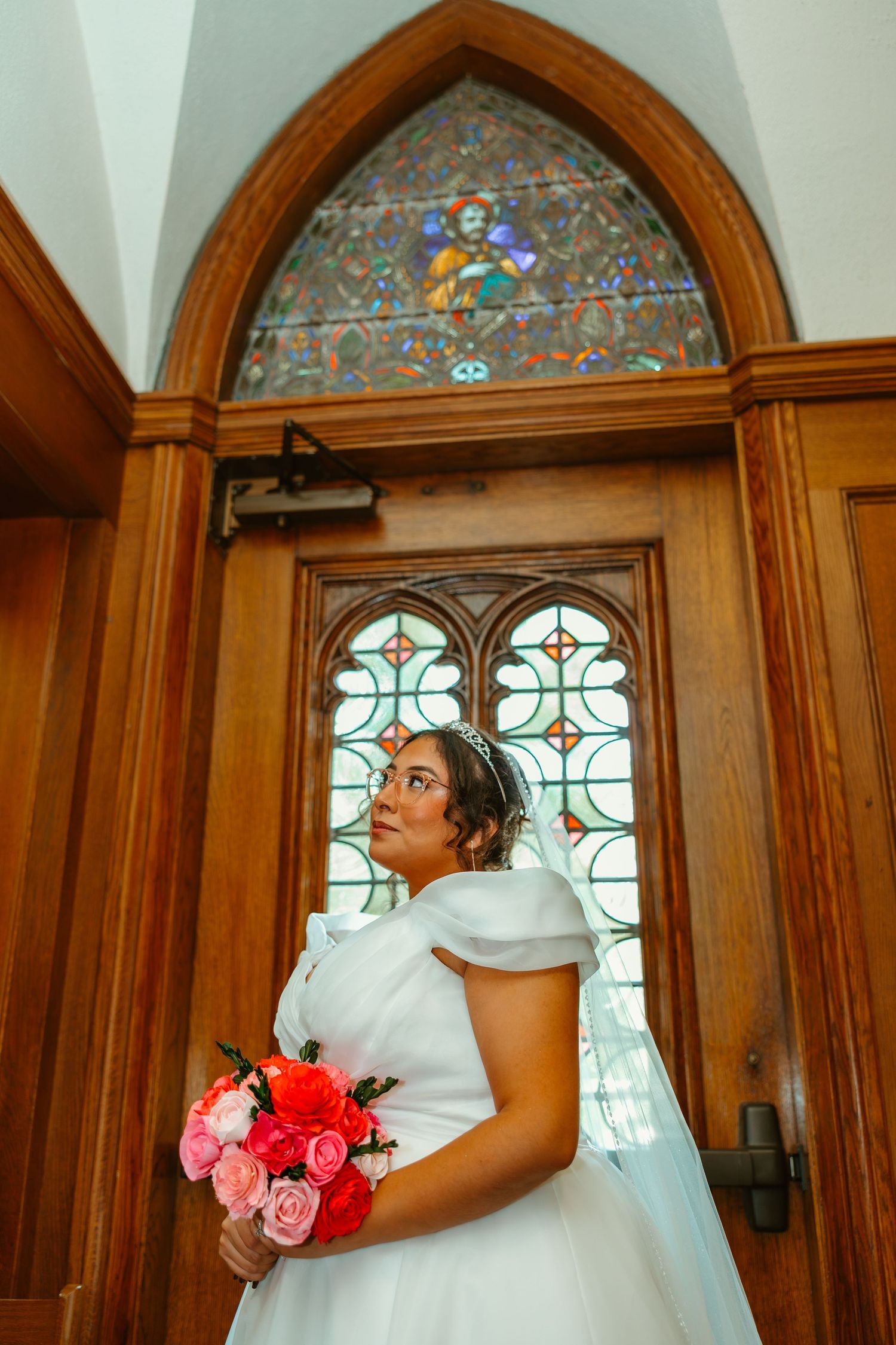 Elegant bride in white off-shoulder gown holding pink bouquet standing before ornate stained glass church door. Chicagoland wedding venues.