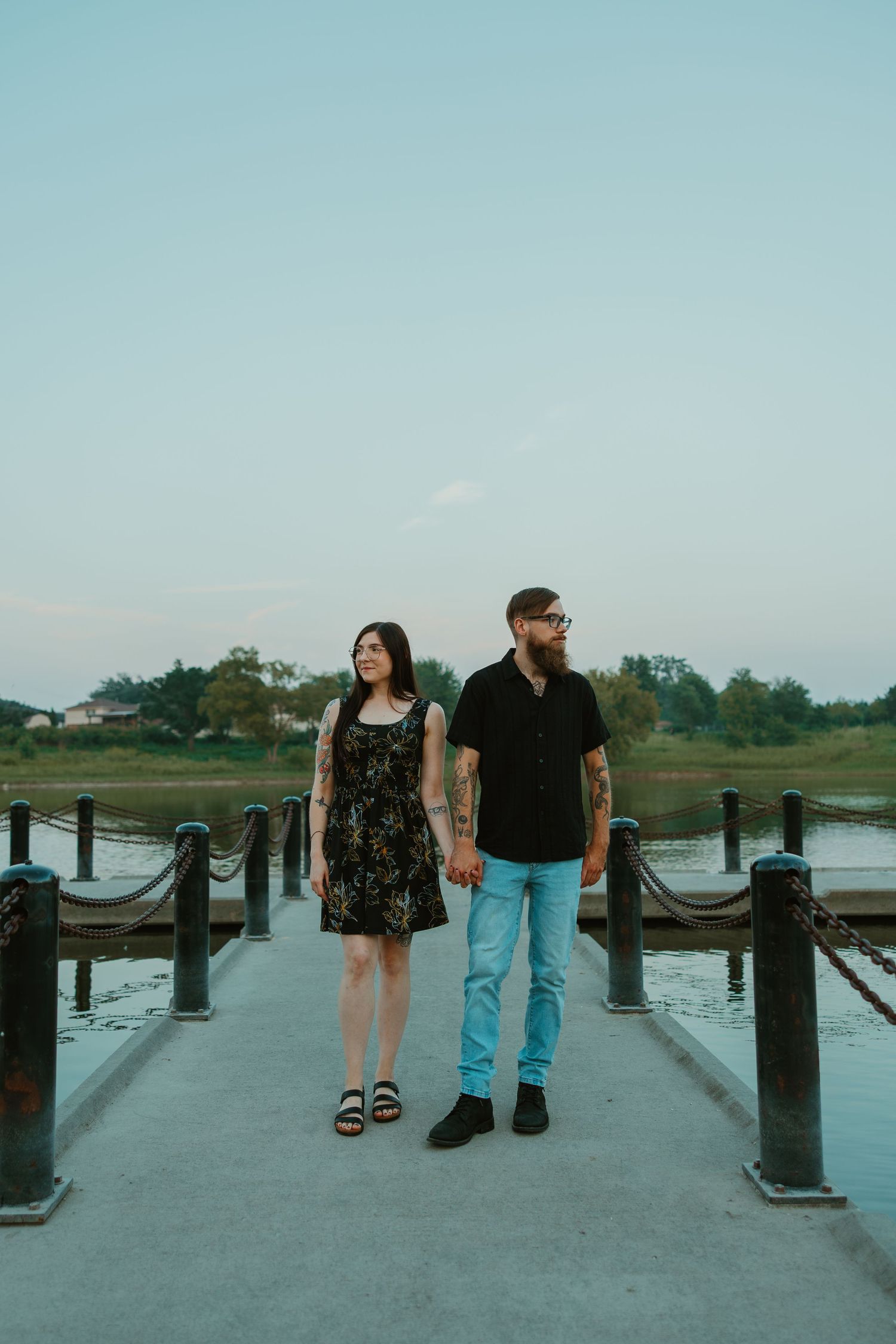 Tattooed couple holding hands on a wooden dock at sunset, standing before a calm lake and clear sky. Chicagoland engagement photos.