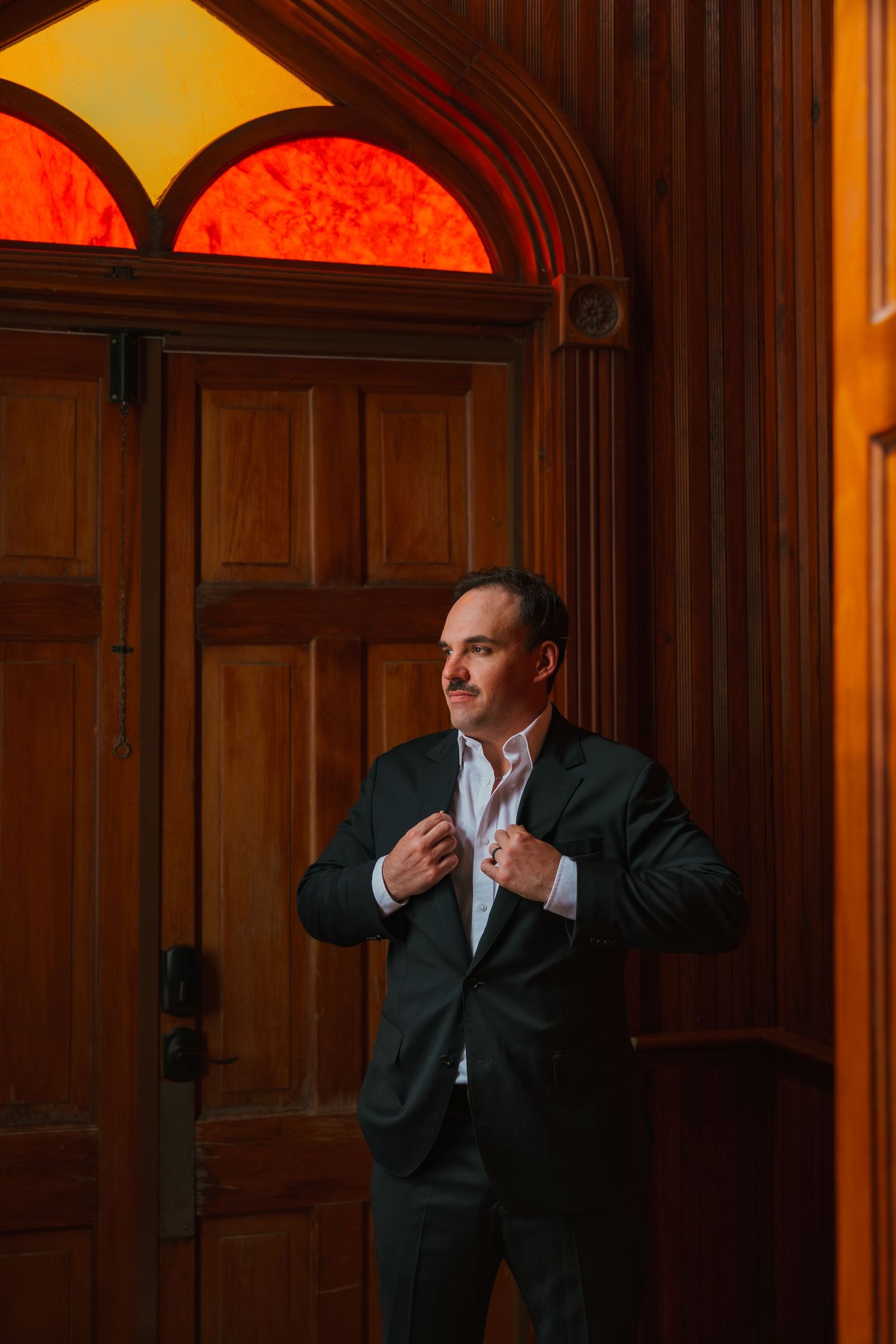 Elegant groom in dark suit adjusting his jacket by ornate wooden doors with amber stained glass. Michigan elopement.