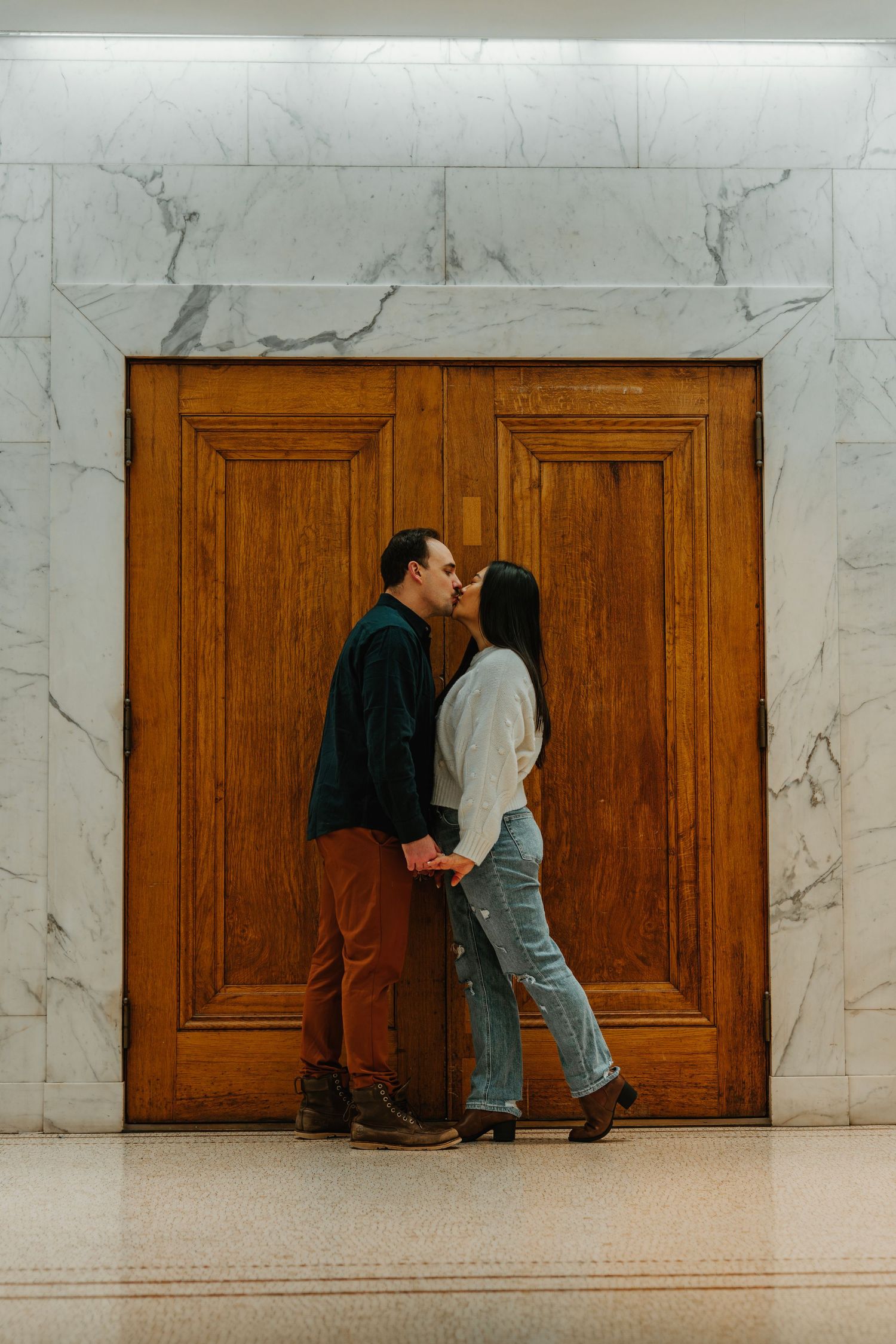 Couple sharing a kiss in front of large wooden doors flanked by white marble walls indoors.