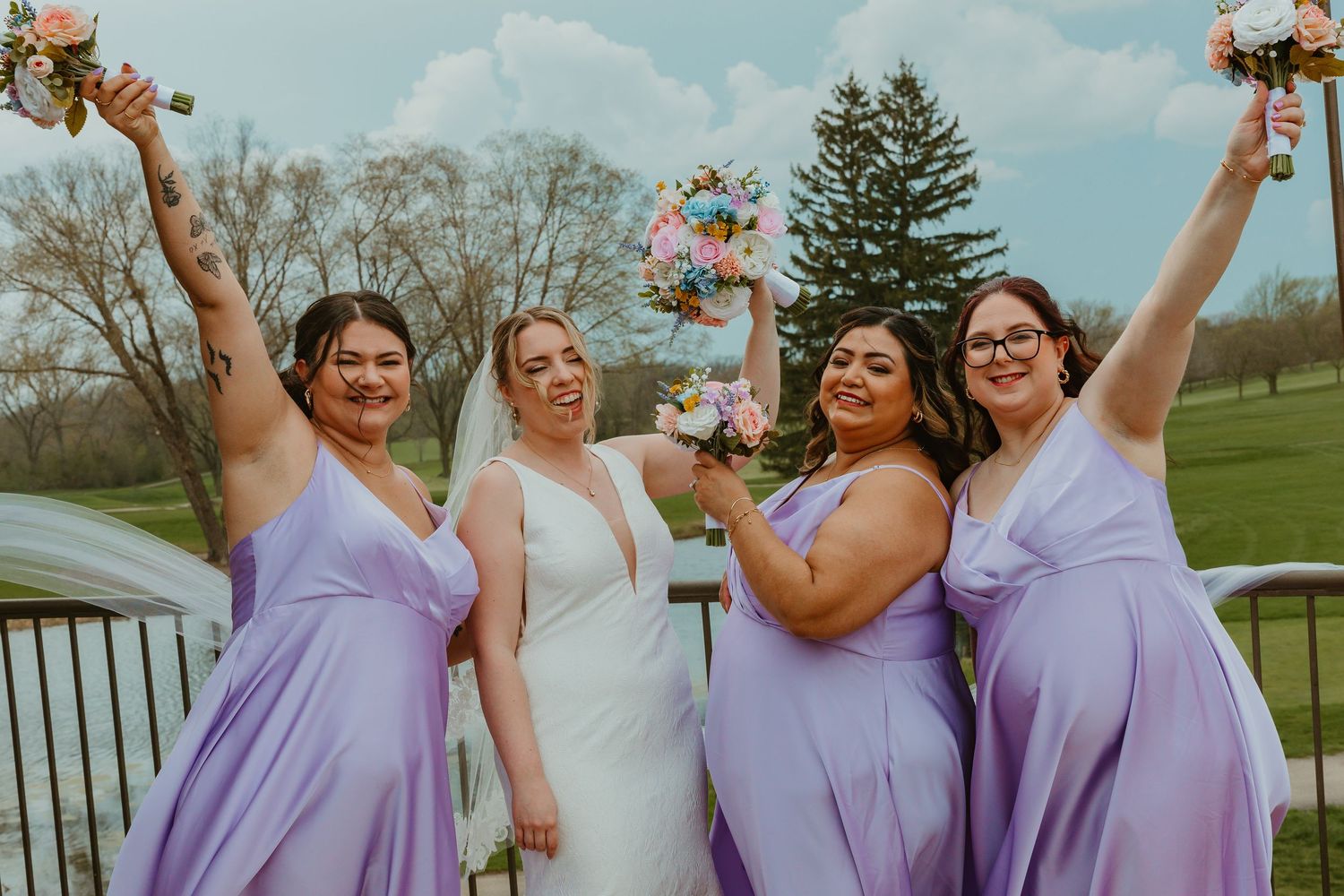Joyful bride and bridesmaids in purple dresses celebrating with raised bouquets outdoors. Chicagoland wedding venues.