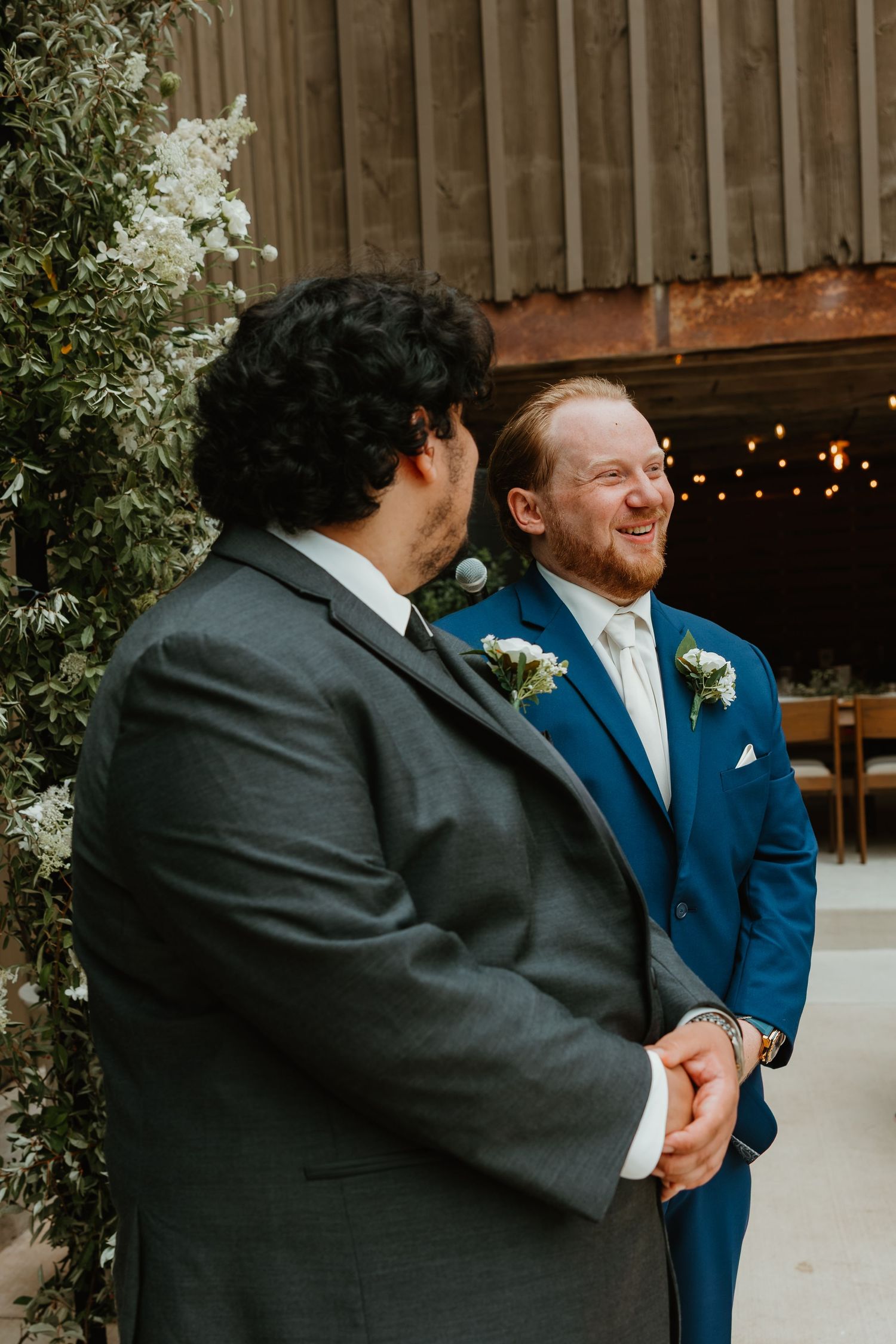 Groom in navy suit laughs joyfully with a groomsman in gray beside a floral arch at a barn wedding. Northwest Indiana wedding venues.