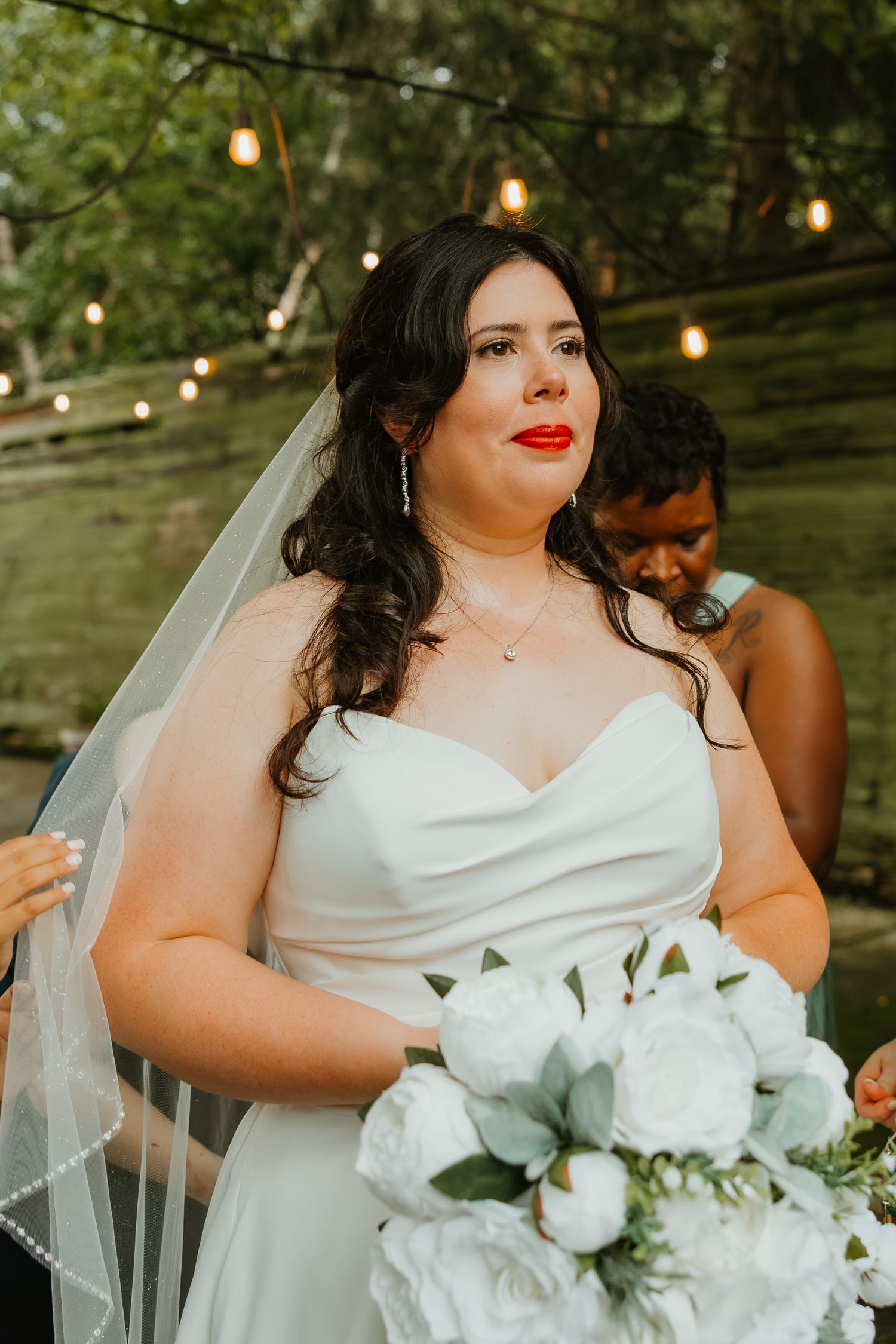 Bride in strapless white gown with veil holds white peony bouquet under string lights outdoors. Northwest Indiana wedding venues.