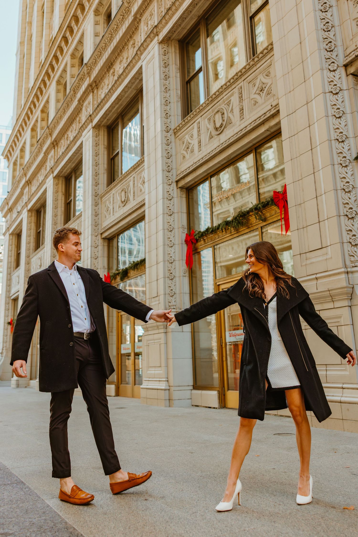 Couple holding hands and smiling at each other on a city sidewalk with a decorated building facade. Outdoor Chicago engagement photos.