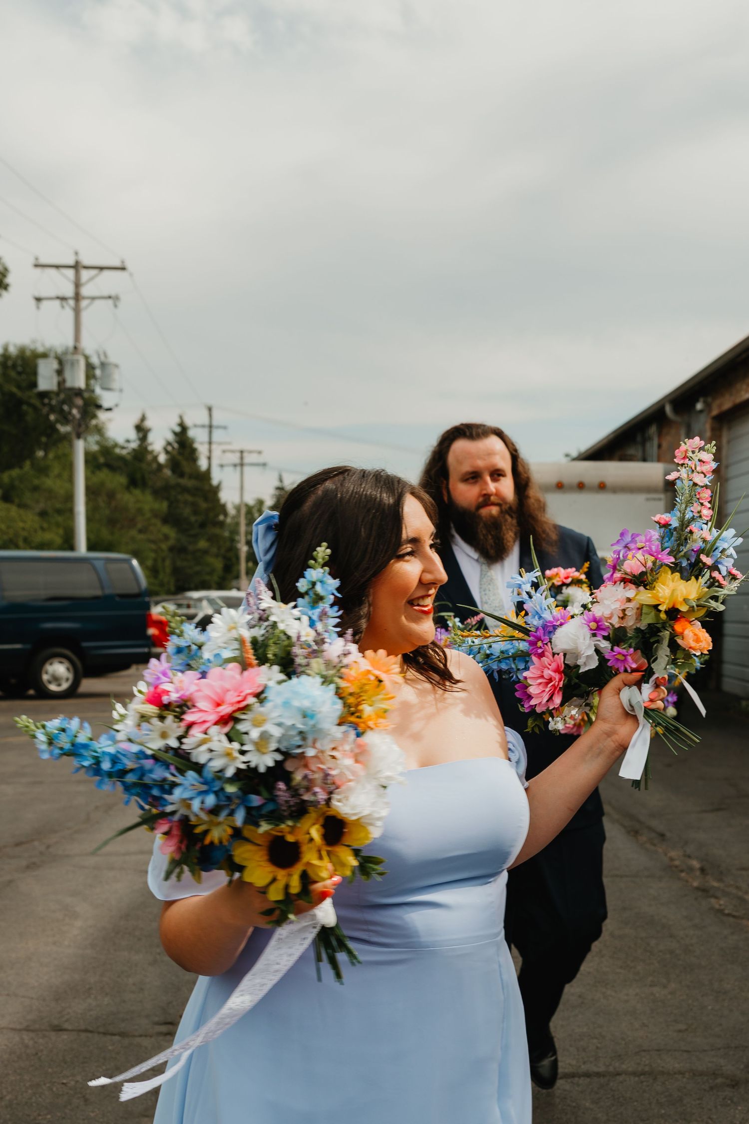 Bride in light blue dress holds colorful bouquet with bearded groom behind her outdoors. Chicagoland wedding venues.