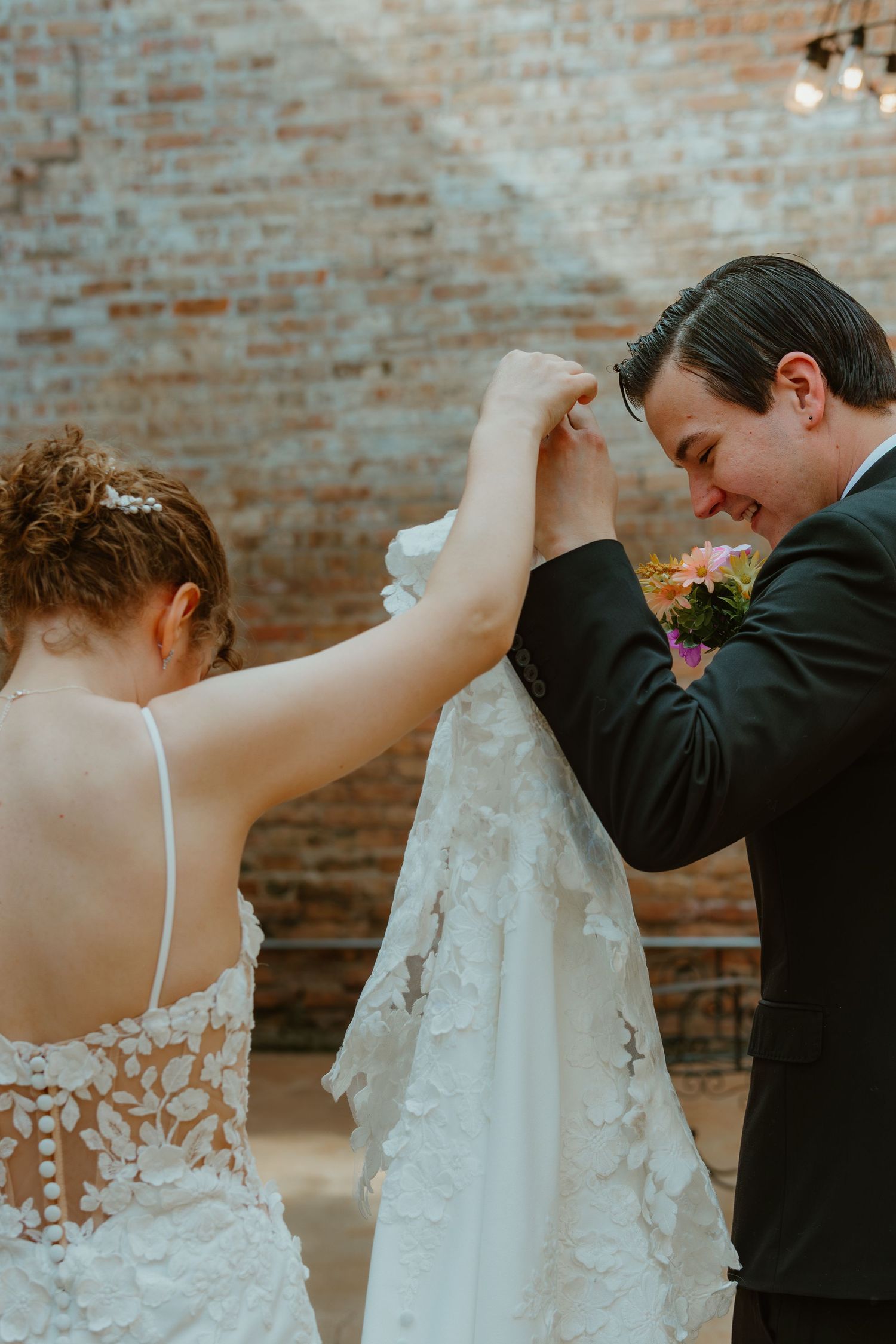 Groom smiling as bride lifts her lace wedding dress train inside a rustic brick-walled venue. Chicagoland wedding venues.