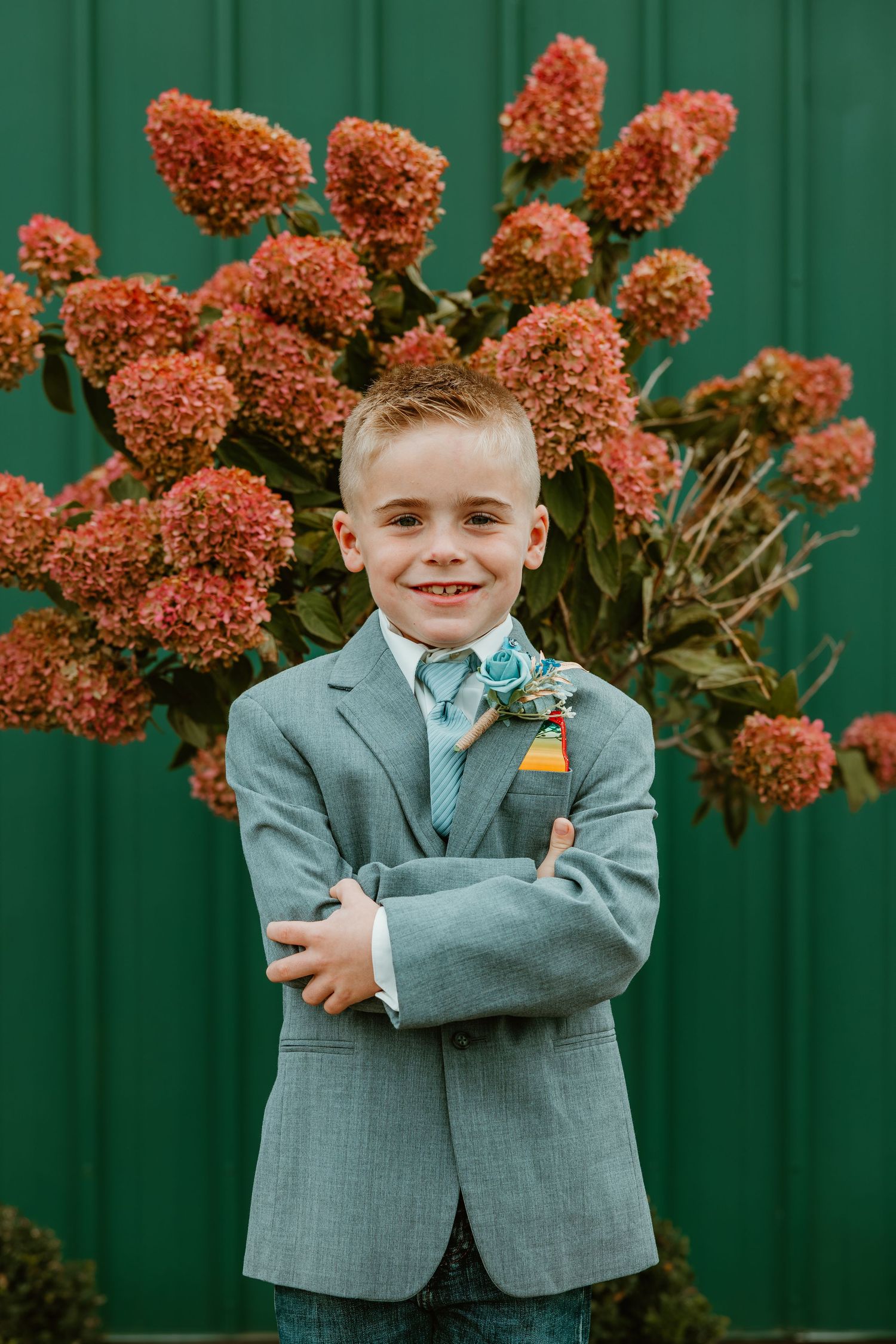 Smiling young ring bearer in gray suit with boutonniere before green wall. Northwest Indiana wedding venues.